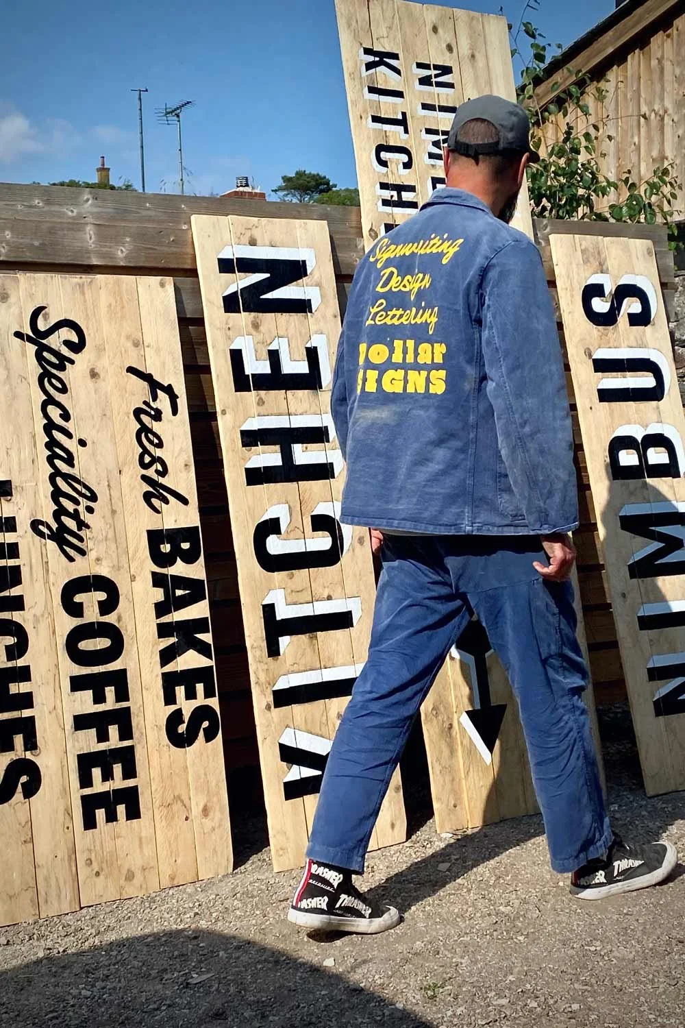 Man in blue vintage French workwear jacket and carpenter pants standing by rustic wooden signs advertising Nimbus Kitchen, Redruth, Cornwall.