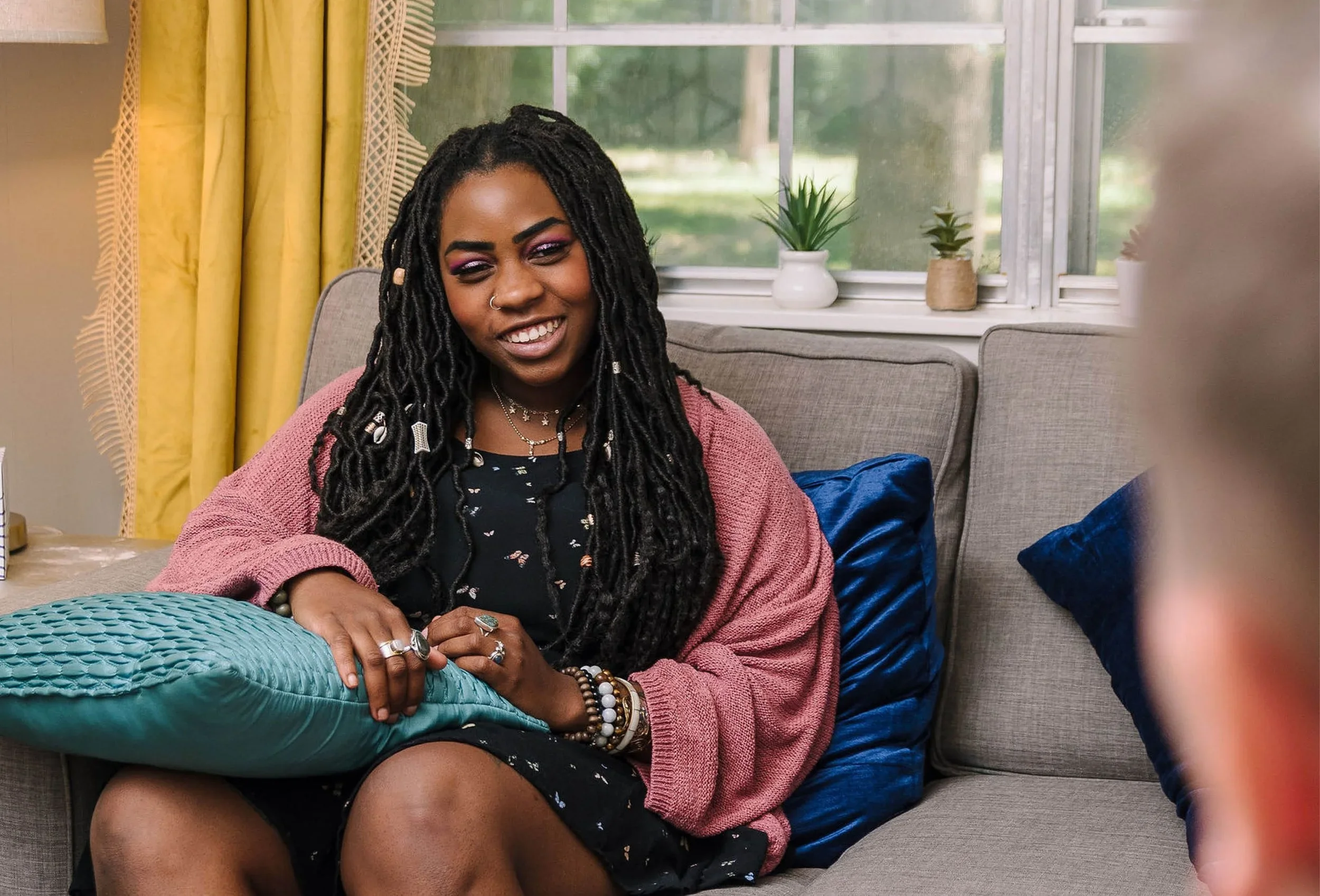 A woman with long dreadlocks, wearing pink eyeshadow and jewelry, sitting on a gray couch in a cozy room with yellow curtains and potted plants on the windowsill, smiling and looking relaxed.