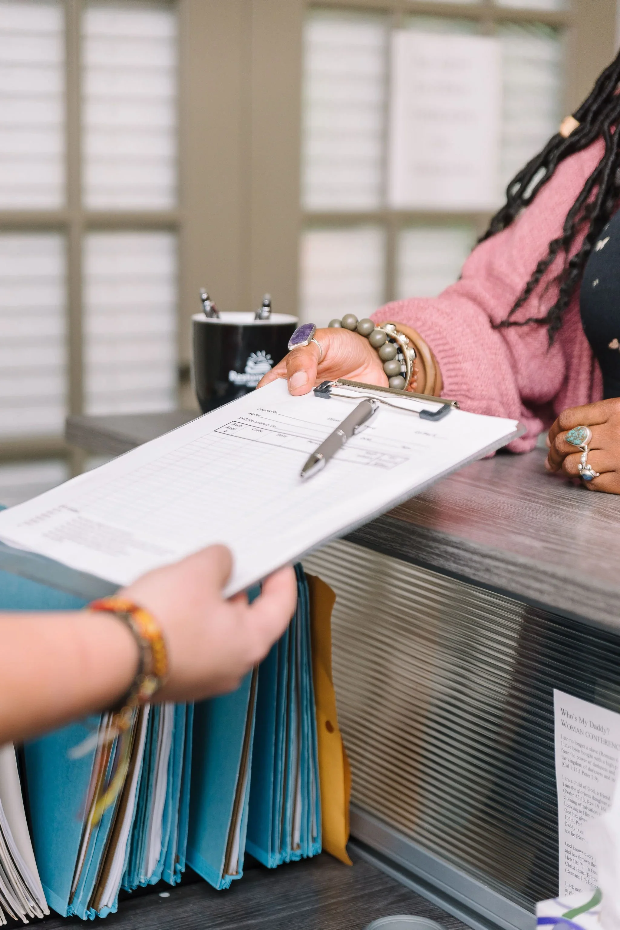 A person's hand holding a clipboard with papers on a desk, with documents and files in the background, some with writing and dates visible.