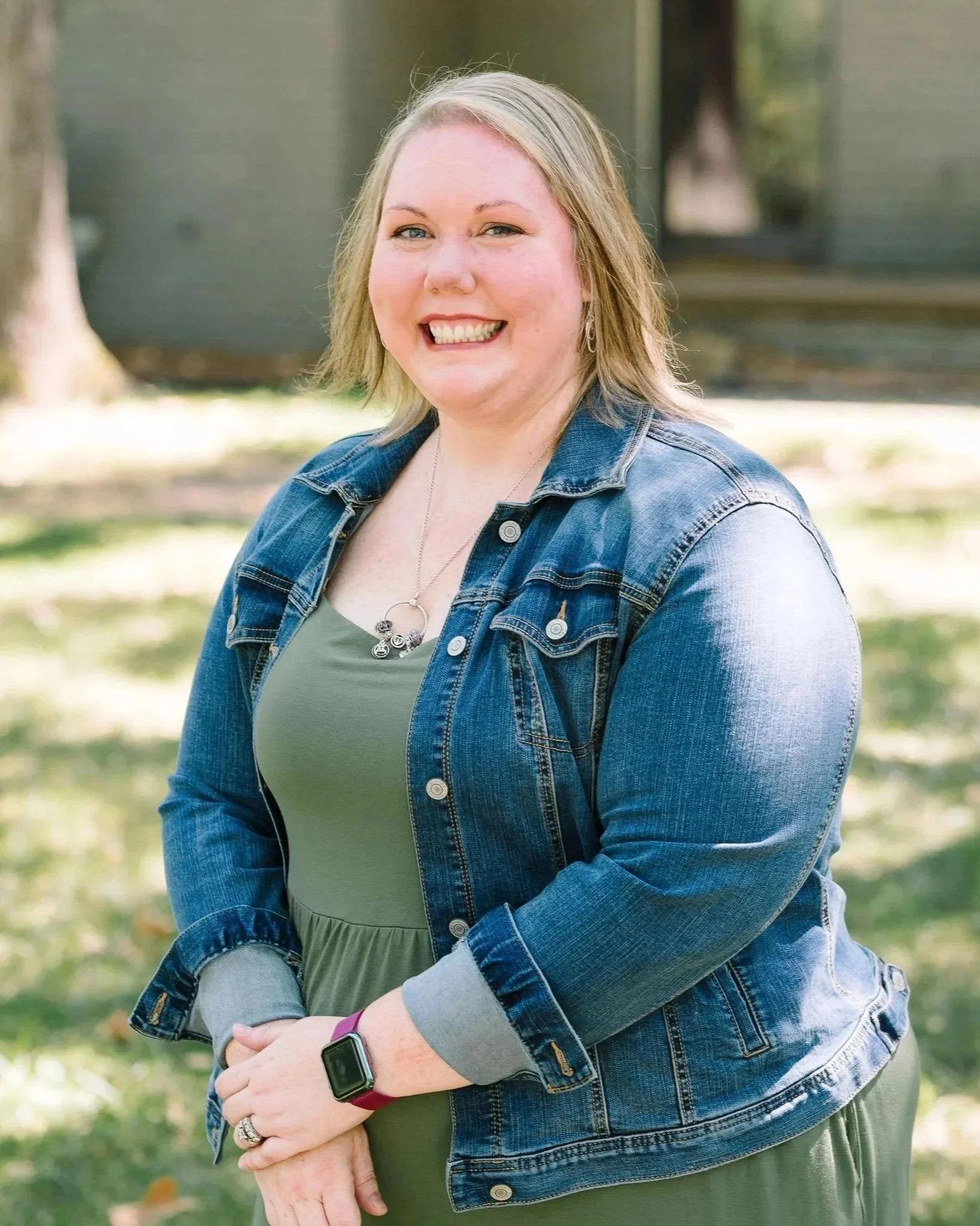 A woman with blonde hair is smiling outdoors, wearing a denim jacket, a silver necklace, and a smartwatch, with trees and blurred background behind her.