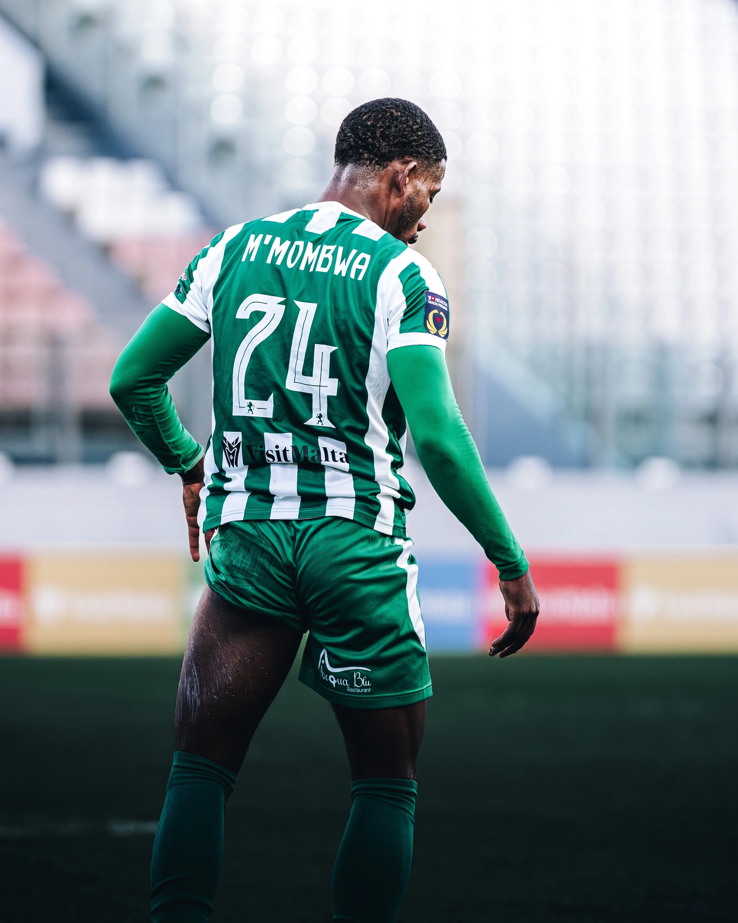 A male football player seen from behind, wearing a green and white striped jersey with the name 'M'MOMBWA' and the number '24' on the back. He is standing on a football field with a blurred stadium background.