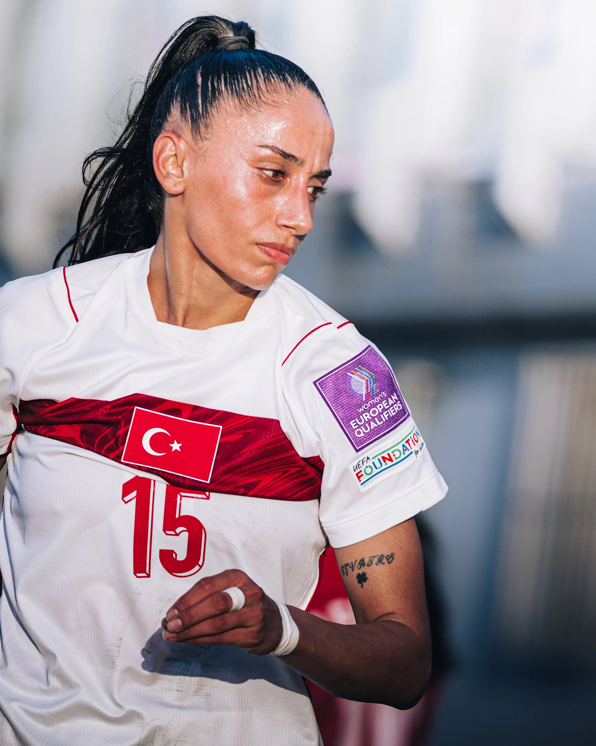 Close-up of a woman wearing a white sports jersey with the flag of Turkey on it, a UEFA Women's European Qualifiers patch, and the number 15. She has dark hair tied in a high ponytail and a tattoo on her left arm with a phrase in script and a four-le