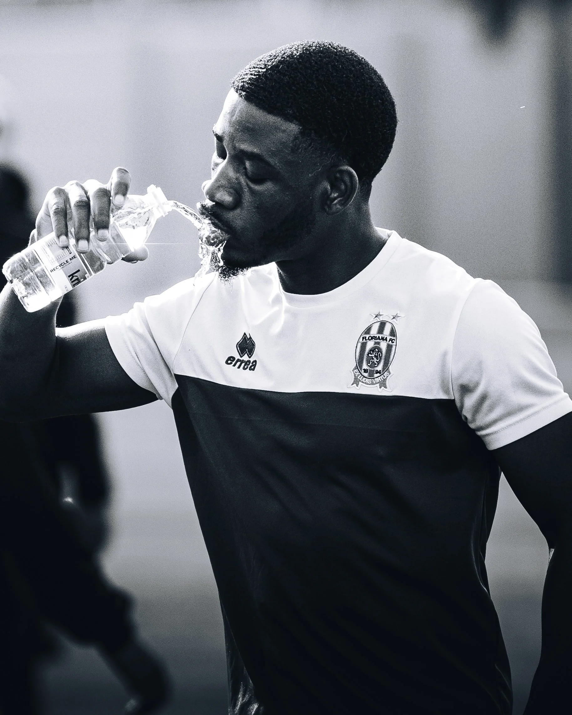 A young man with short hair, a beard, and wearing a soccer jersey with a Floriana logo, is drinking water from a plastic bottle.