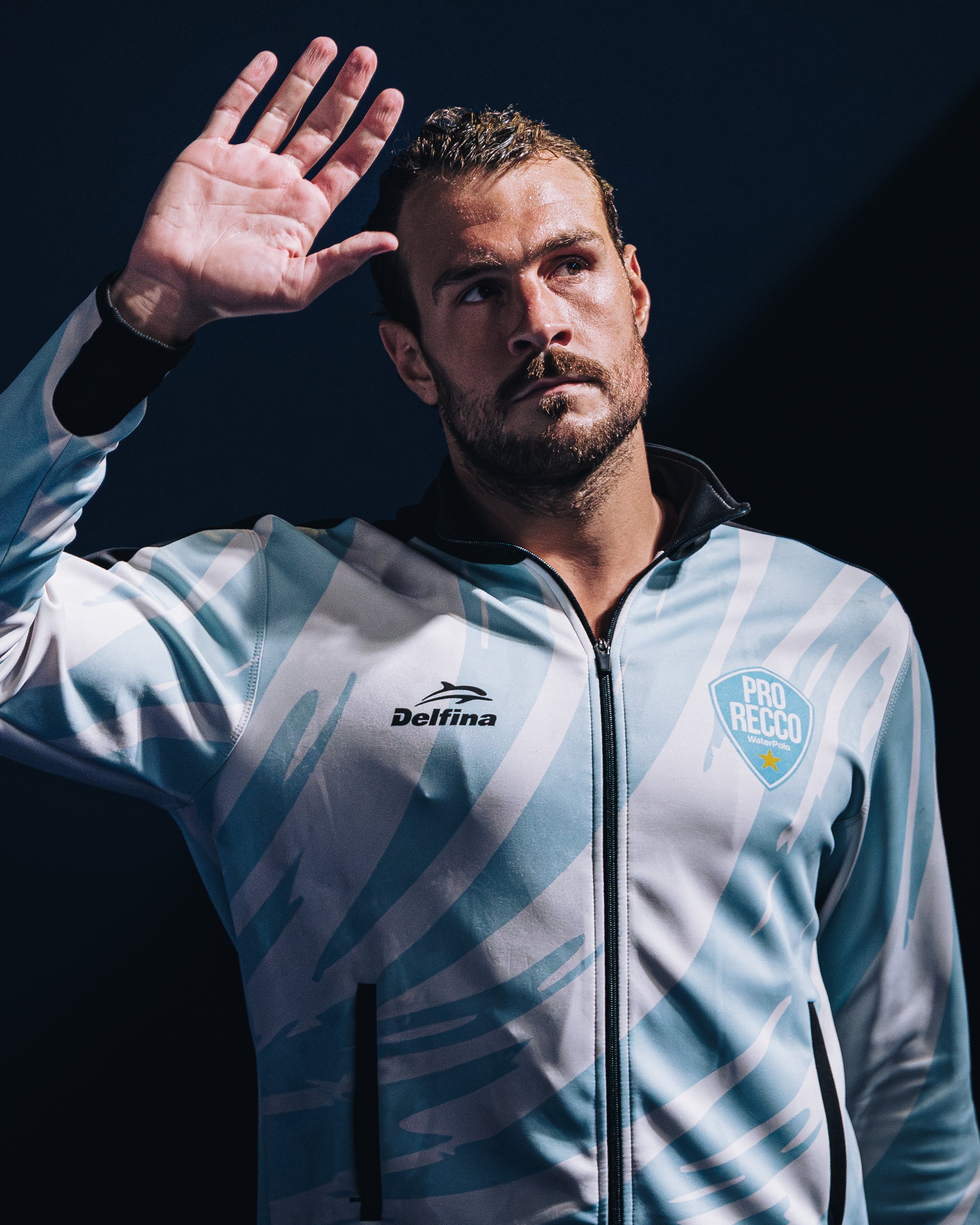 A male athlete raising his right hand in a gesture of greeting or acknowledgment, wearing a sports jacket with logos, with a dark background.