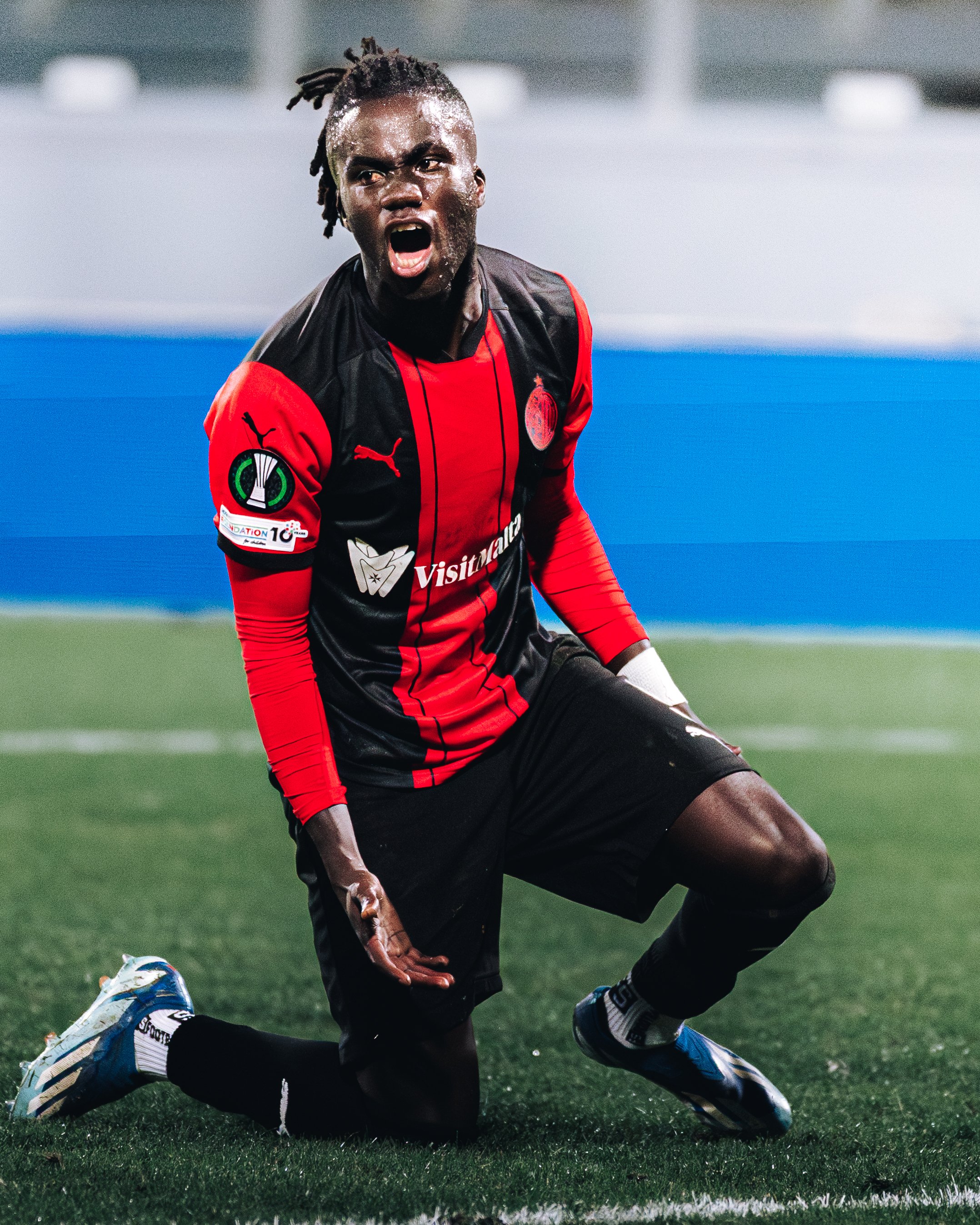 A football player with dreadlocks and dark skin, wearing a black and red jersey, is kneeling and shouting on a football field. The player appears to be in a moment of intense emotion during a game.