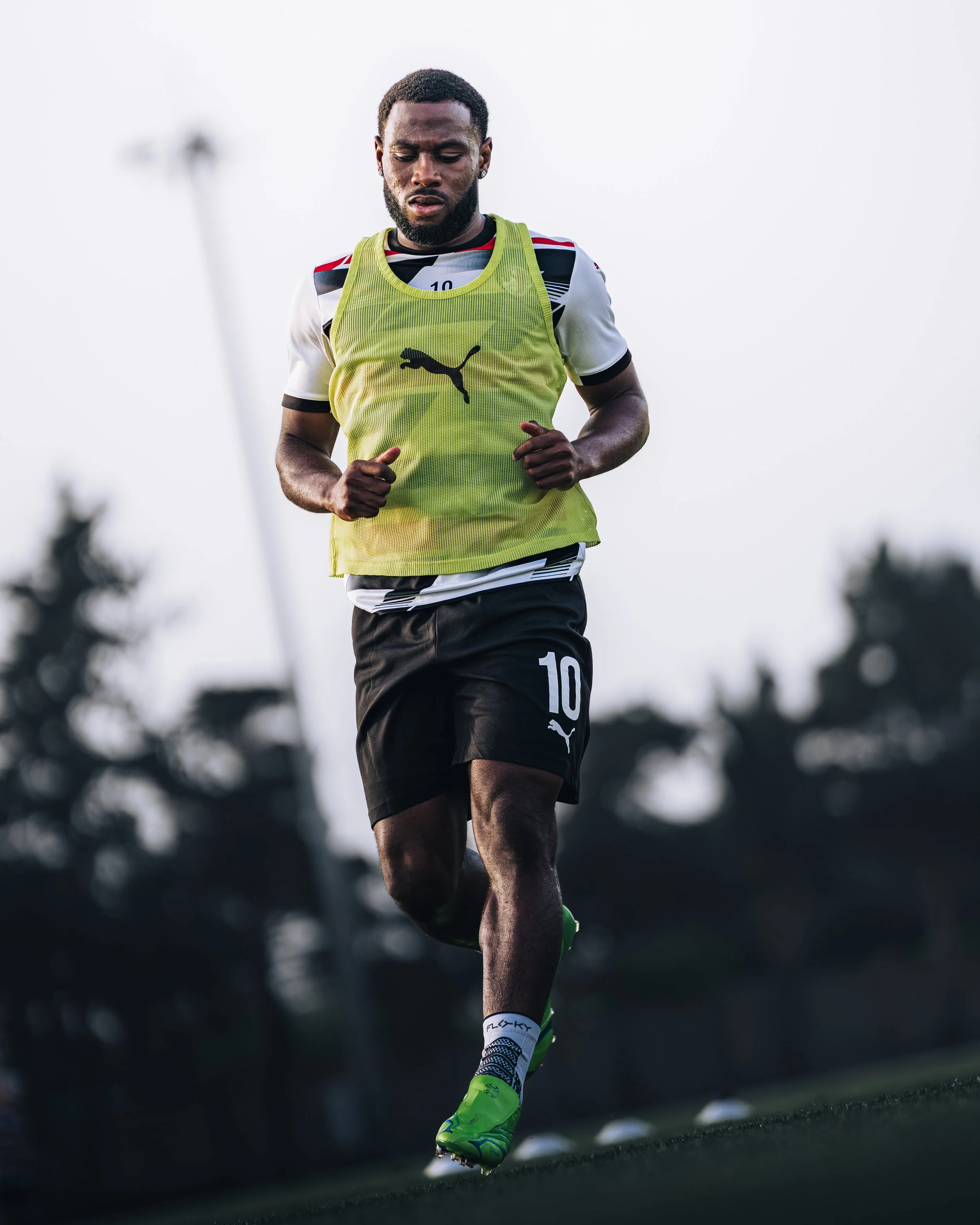 A male athlete running on a track, wearing a yellow Puma sports jersey, black shorts with the number 10, neon green cleats, and black socks, with blurred trees in the background.