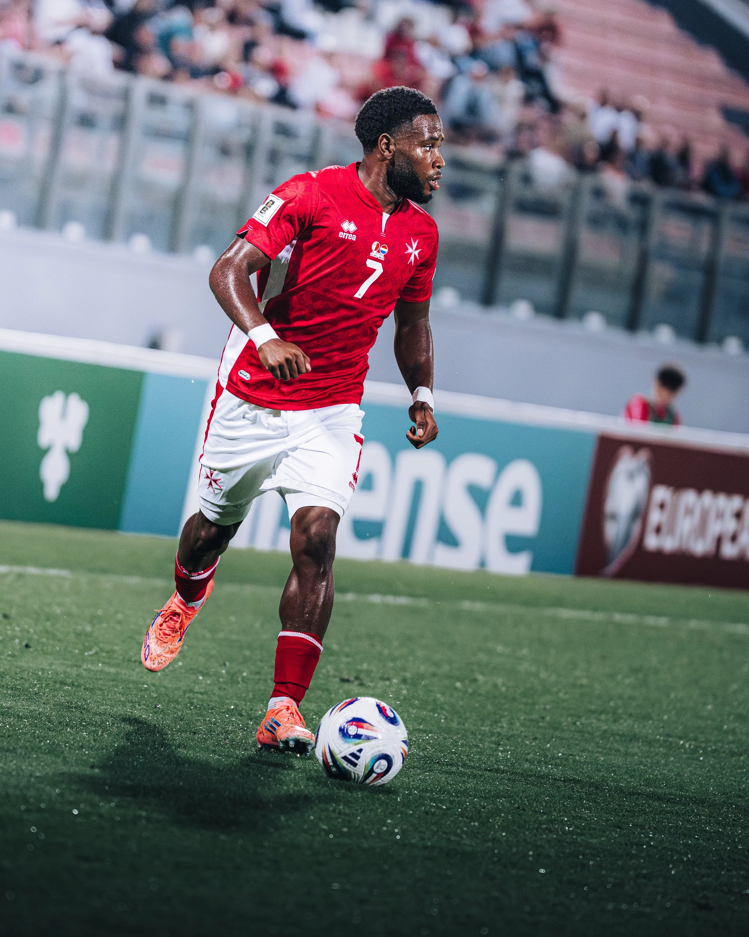 A soccer player wearing a red jersey with the number 7 is running on the field, controlling the ball with his right foot. The crowd is blurred in the background.