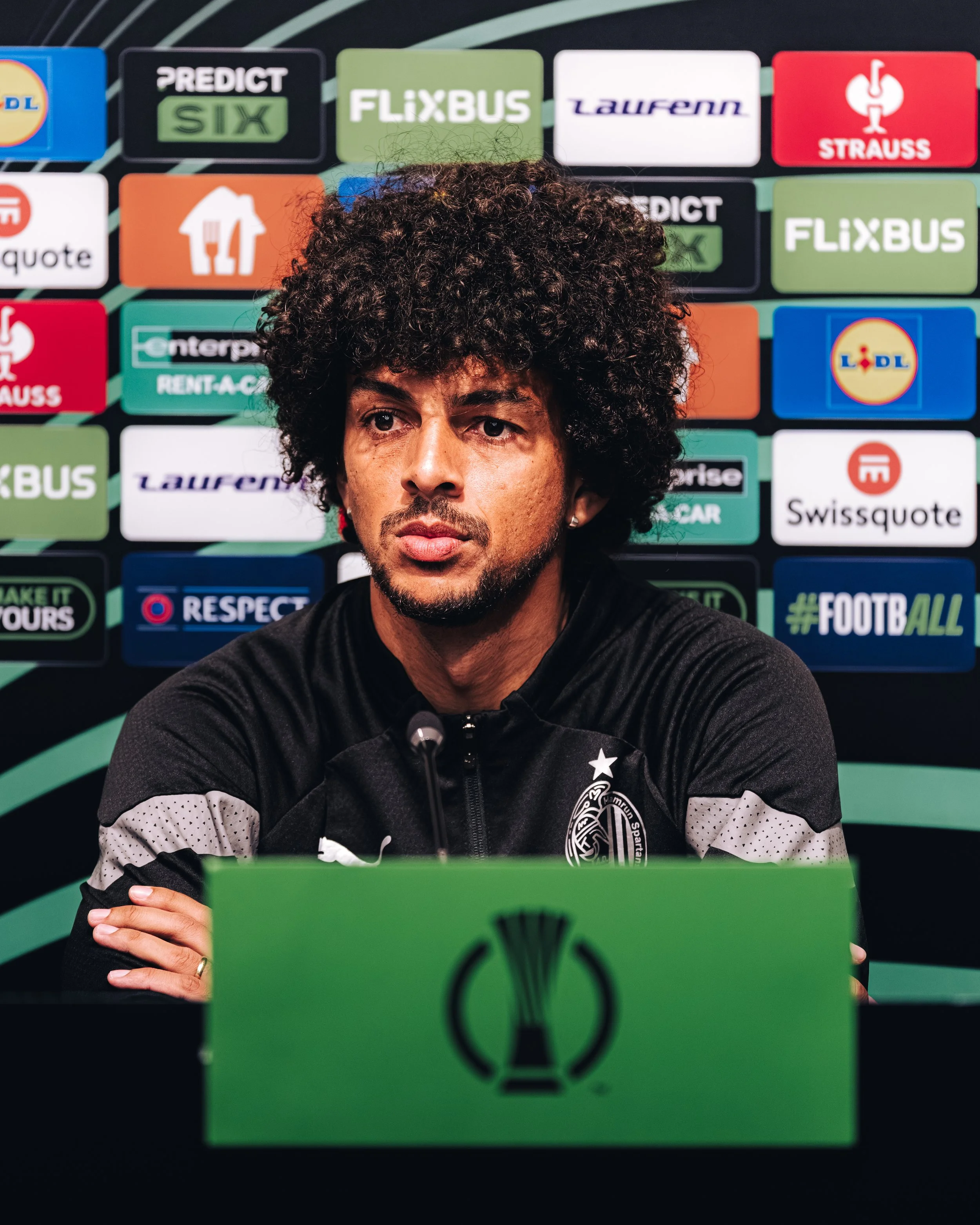 An athlete, Marcelina, Hamrun's centreback at UEFA Conference League press conference.  The background has logos of various brands including Lidl, Flixbus, Laurenn, Strauss, Swissquote, and others, indicating a press conference or media event.