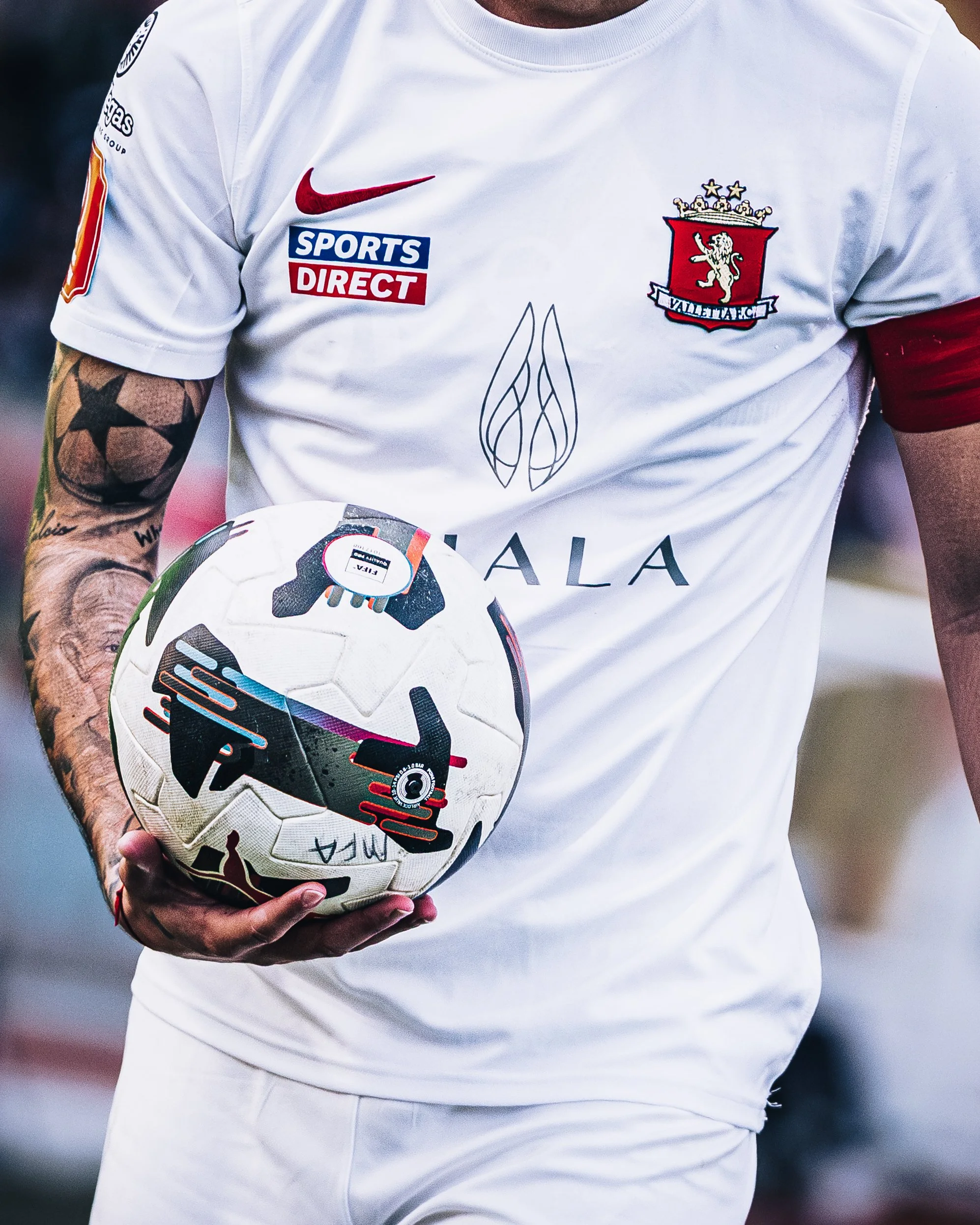 Close-up of a person wearing a white sports jersey with various logos, holding a soccer ball, showing part of their arm and chest.