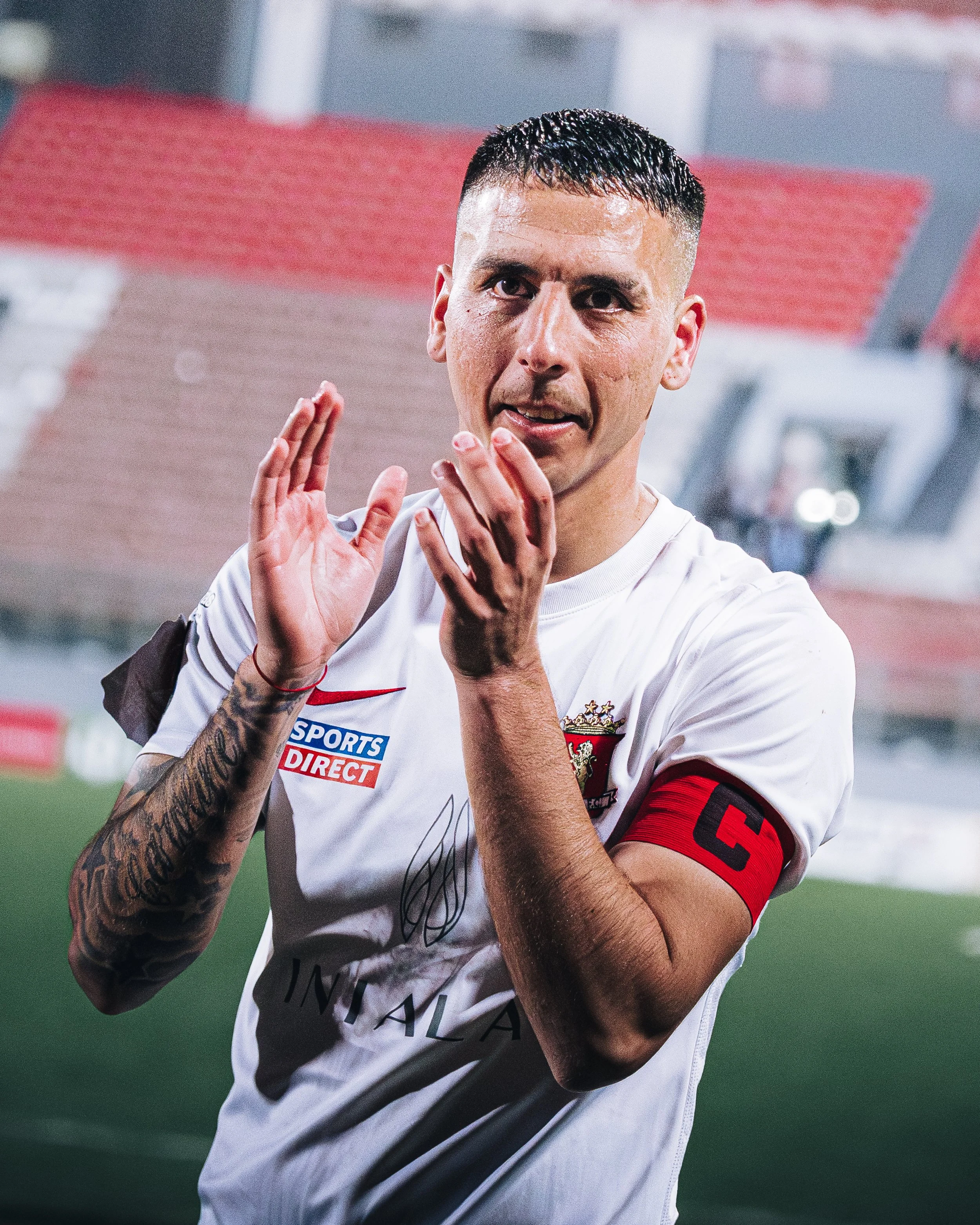 A soccer player on the field wearing a white jersey with red and black details, a red captain's armband, and making a gesture with his hands. He has short black hair and visible tattoos on his left arm.