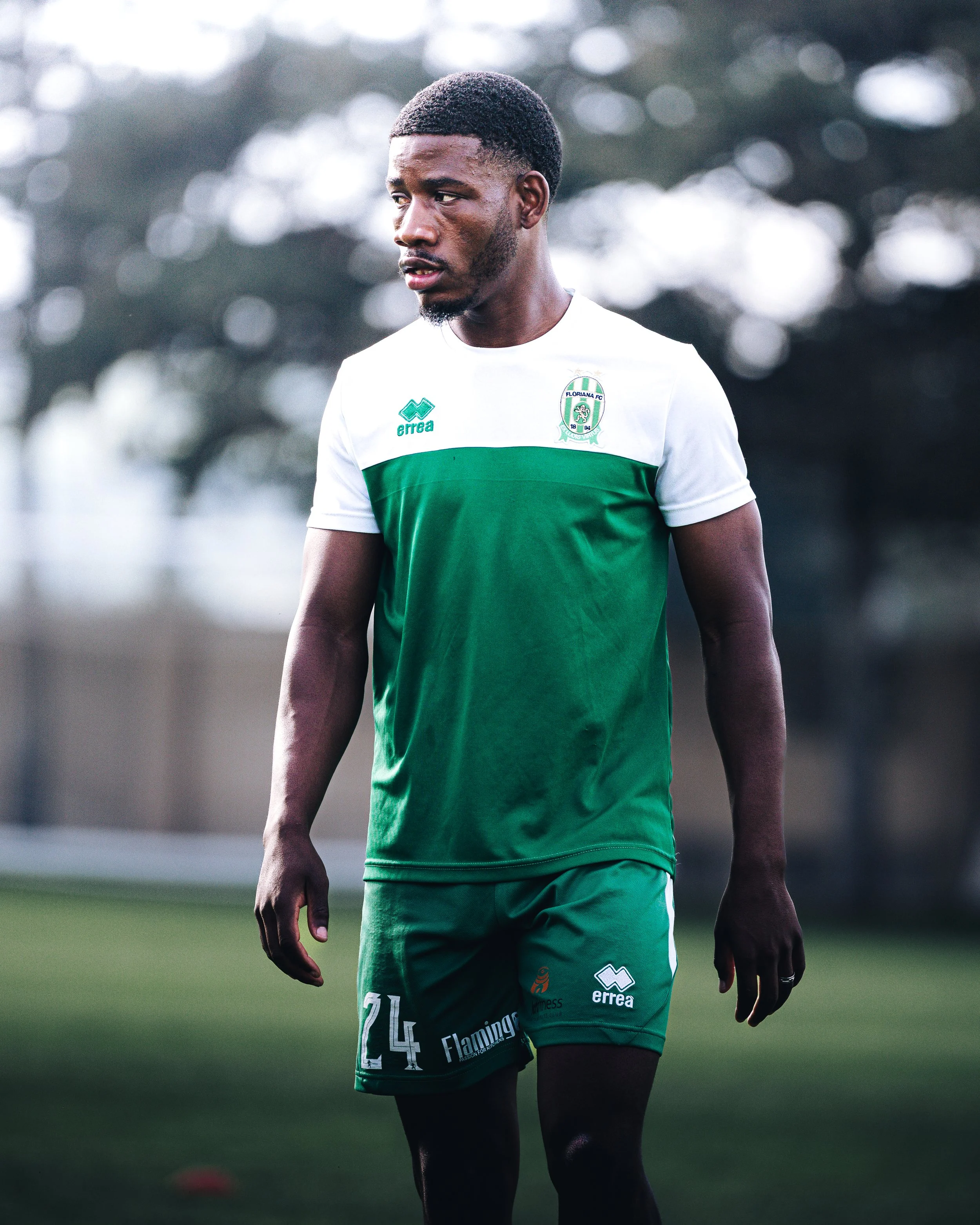 A male football player with dark skin and short hair, wearing a green and white jersey with the number 24. He is on a football field with a blurred background of trees, looking to his left with a focused expression.