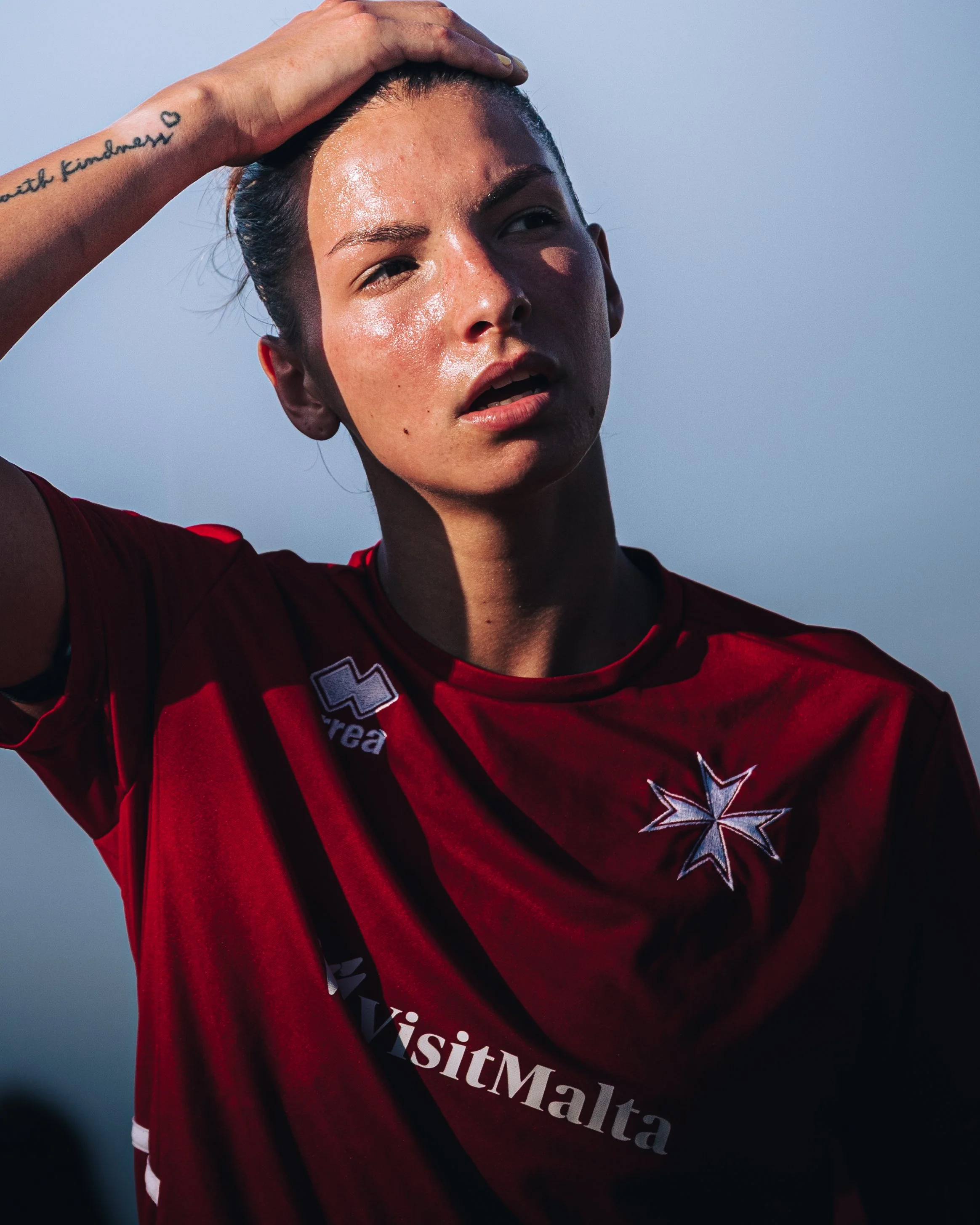 Close-up of a woman with her hand on her forehead, looking into the distance, wearing a red sports jersey with white and black text, and a team logo. Her skin is shiny and sweaty, suggesting she just finished a sport activity.