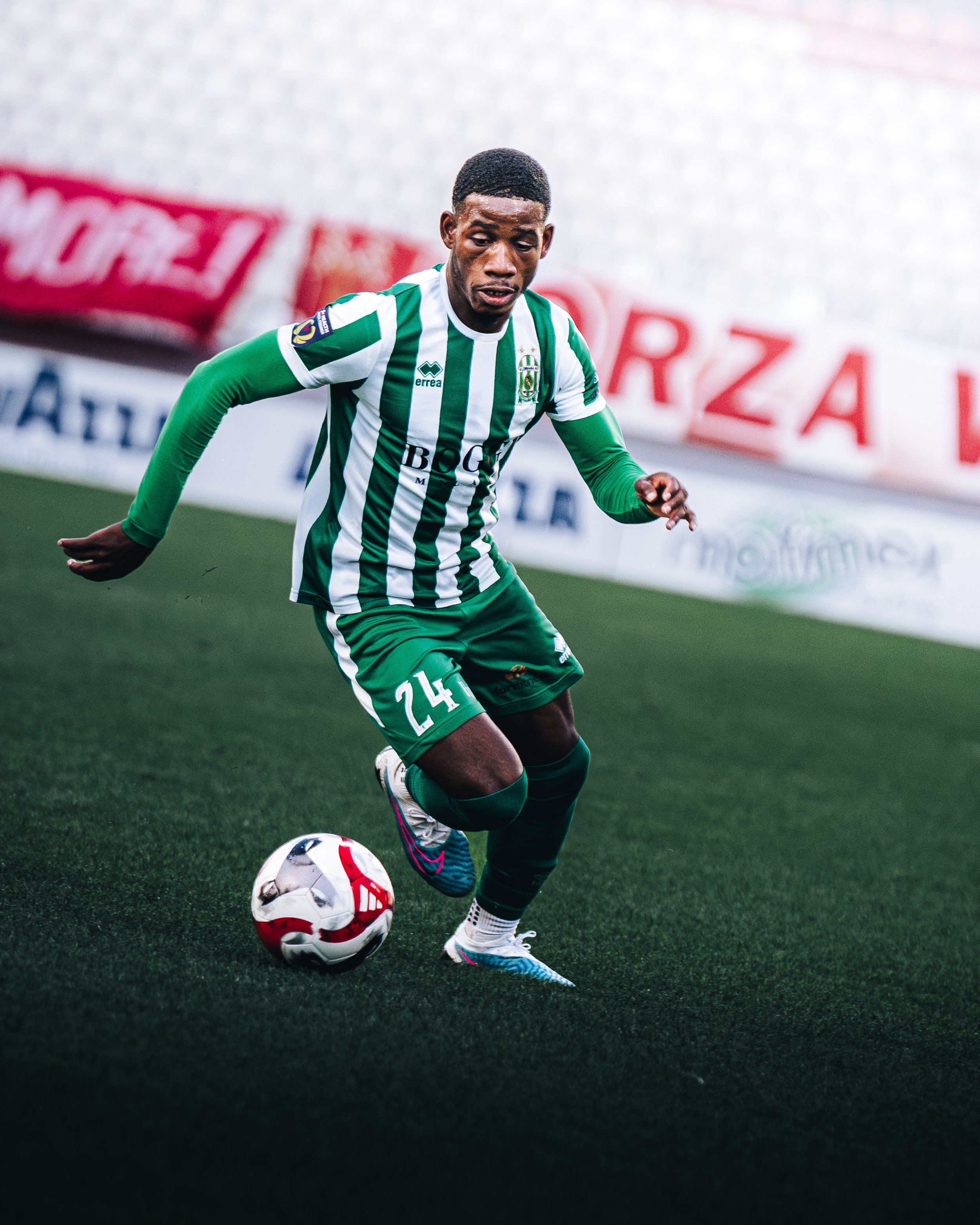 A soccer player dressed in green and white striped uniform with the number 24 on shorts, dribbling a soccer ball on a green field.