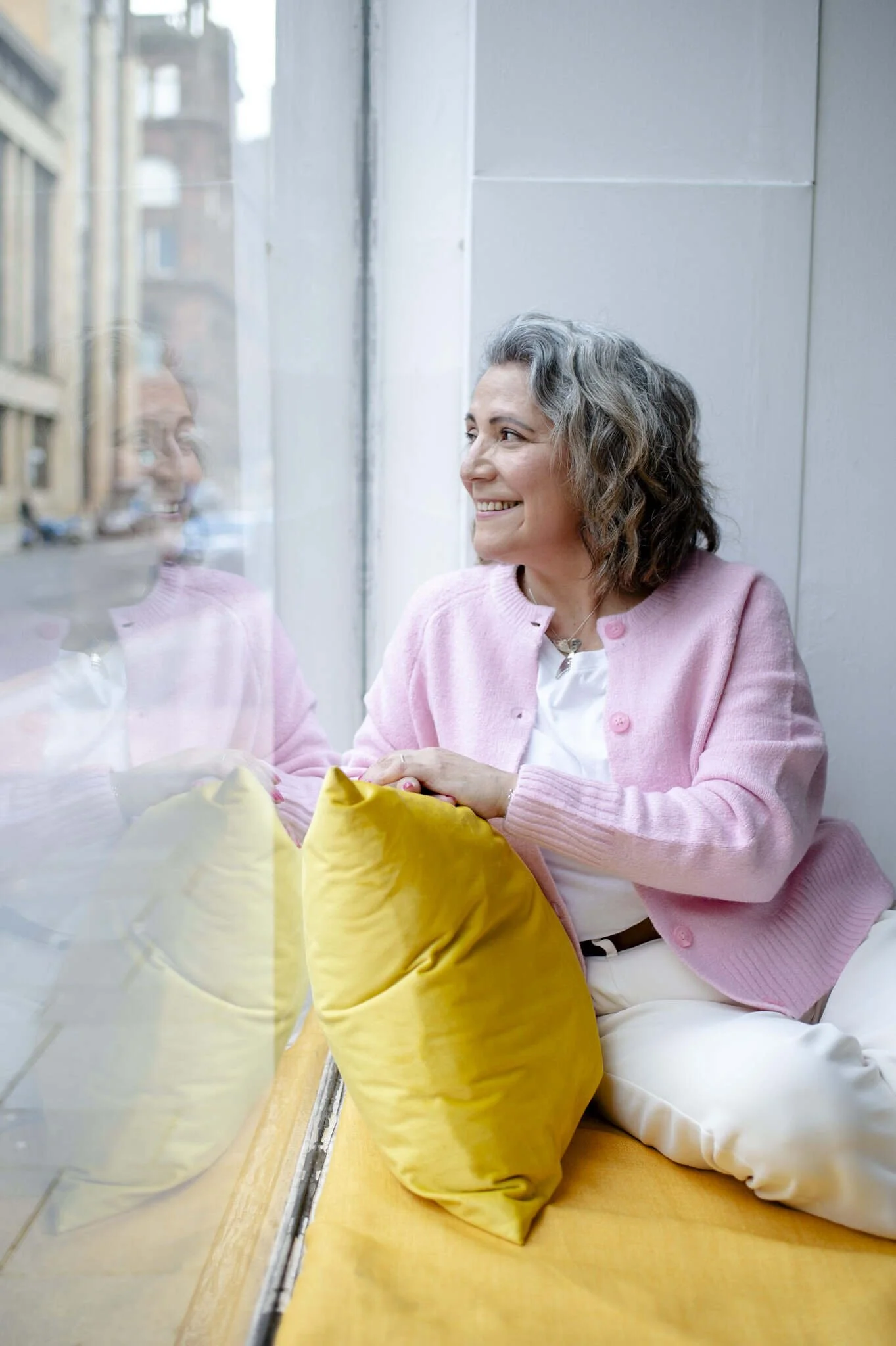 A woman with grey curly hair wearing a pink cardigan and white trousers sitting by a window, smiling while sitting on a a yellow velvet sofa, looking outside.