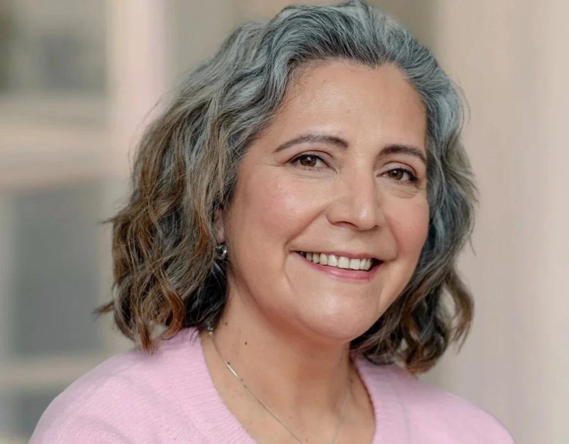 A woman with shoulder-length wavy gray hair, smiling, wearing a pink top and a necklace, indoors.