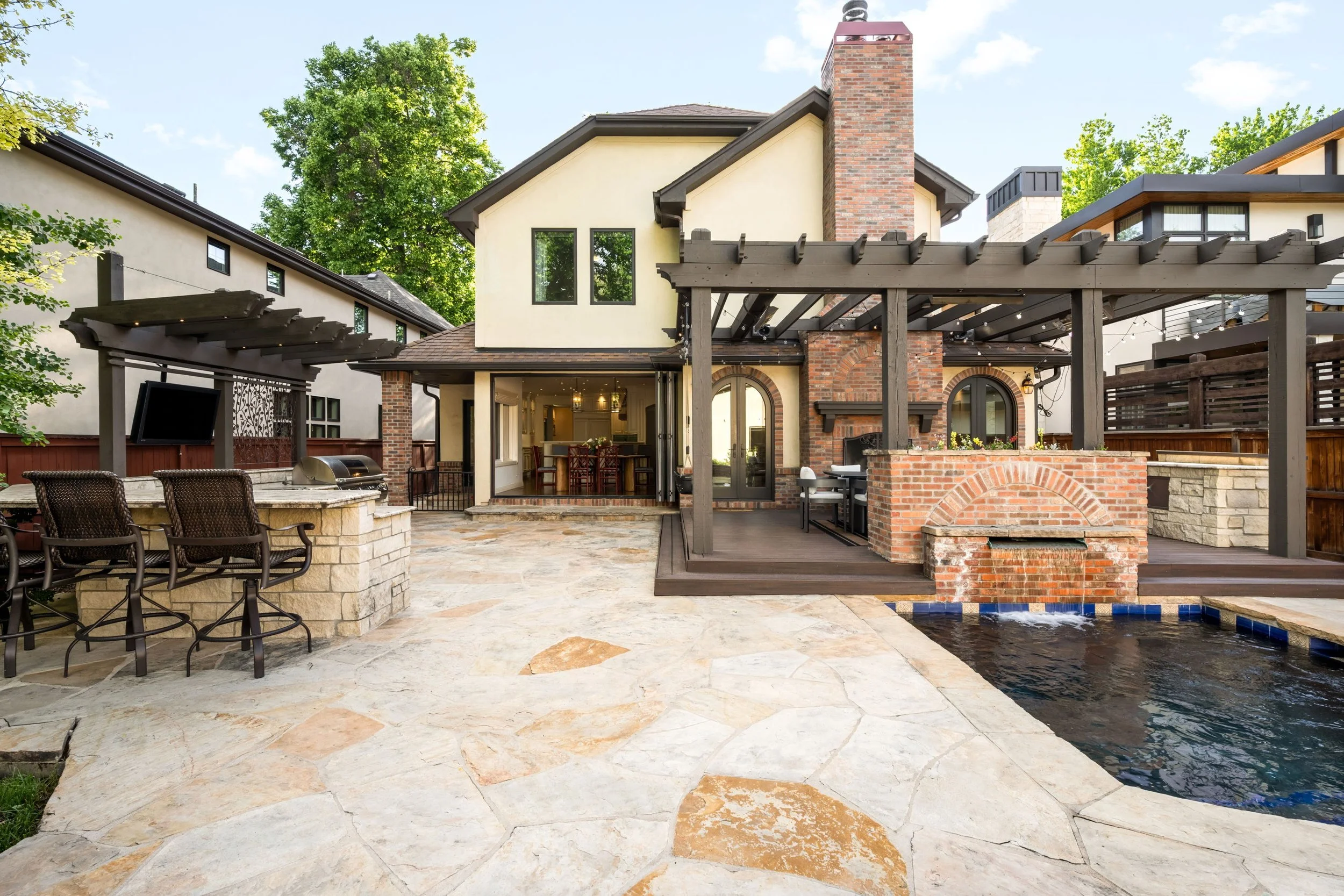 Backyard with a stone patio, pool, outdoor fireplace, wooden pergola, and neighboring houses with trees in the background.