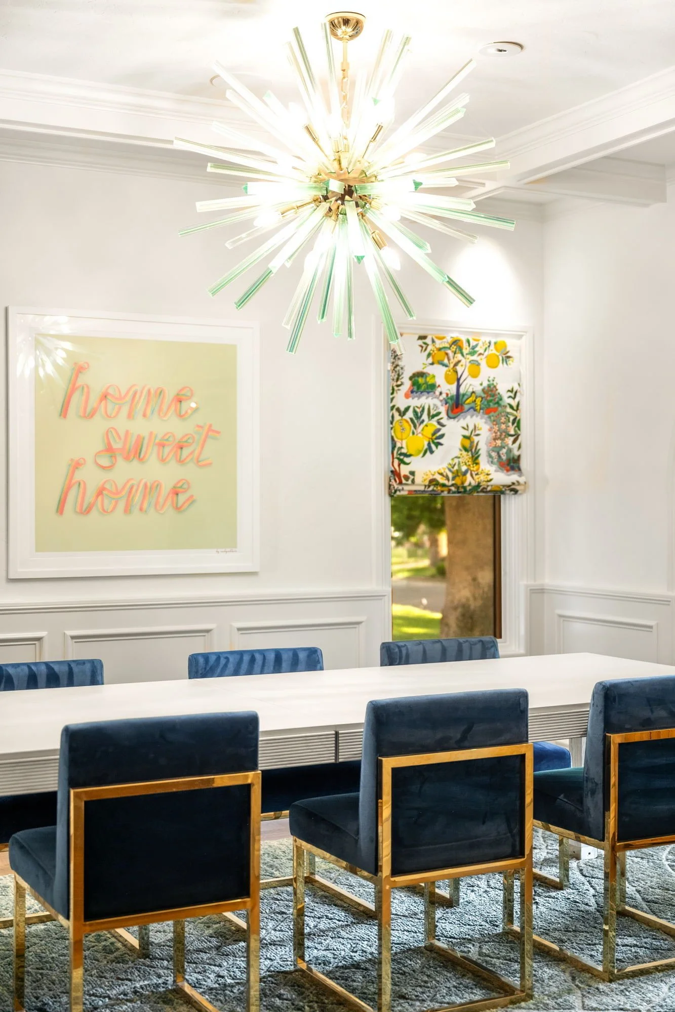 Modern dining room with a white table, navy blue chairs with gold frames, a statement chandelier, and decorative wall art featuring the phrase 'home sweet home' and colorful lemon-themed artwork. A window with a patterned roman shade shows a view of 