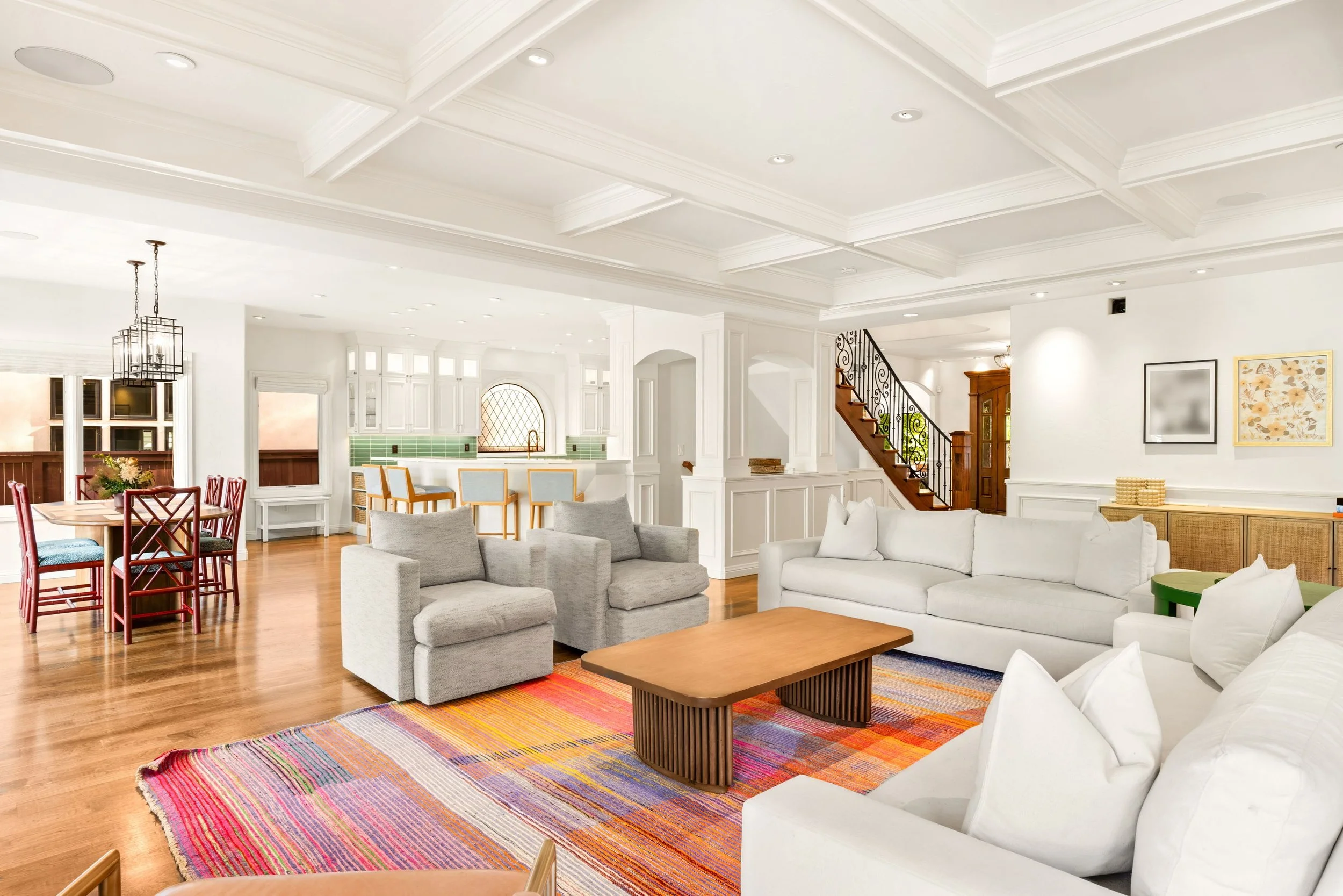 Bright and spacious living room with white walls and ceiling, hardwood floors, a colorful striped rug, multiple sofas, a wooden coffee table, and a dining area with red chairs. The kitchen in the background features white cabinets, green backsplash t