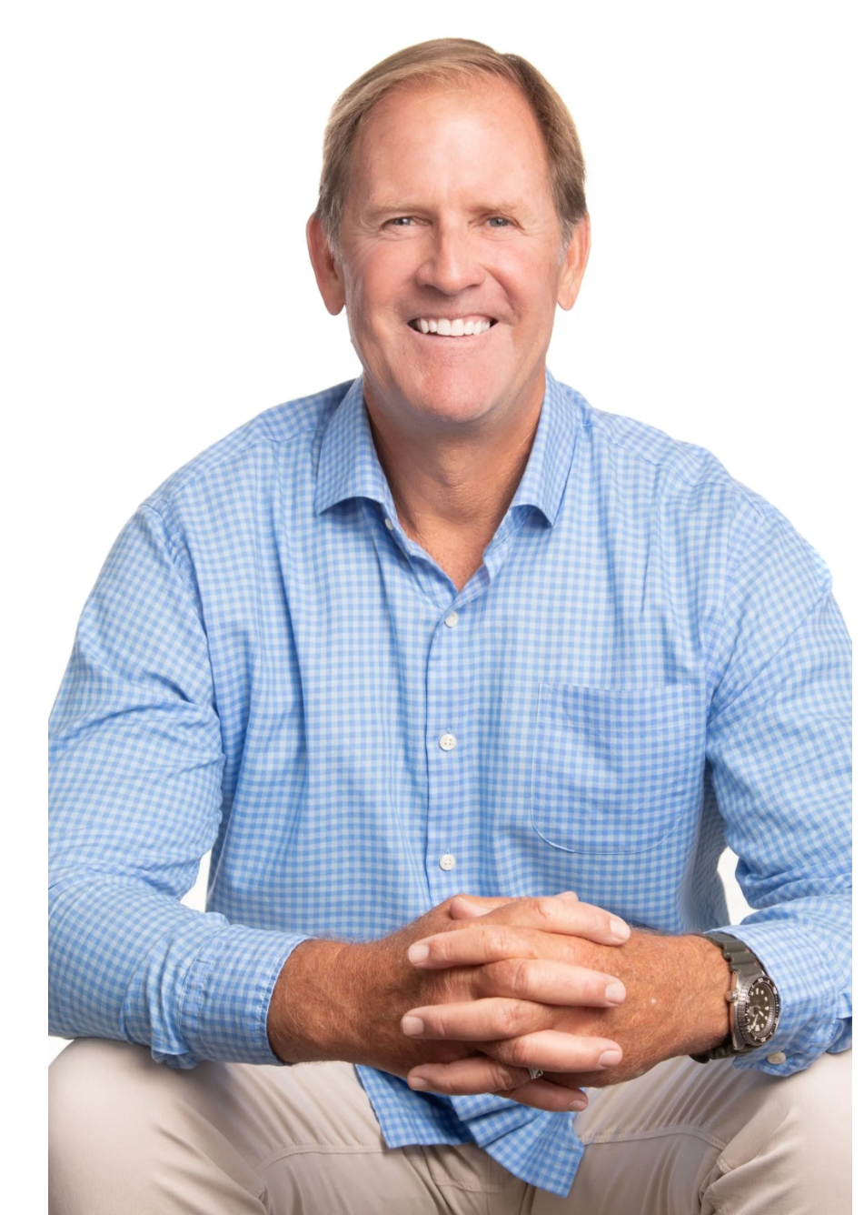Portrait of a smiling middle-aged man with light brown hair, wearing a blue checkered button-up shirt and a wristwatch, sitting with hands clasped together against a white background.
