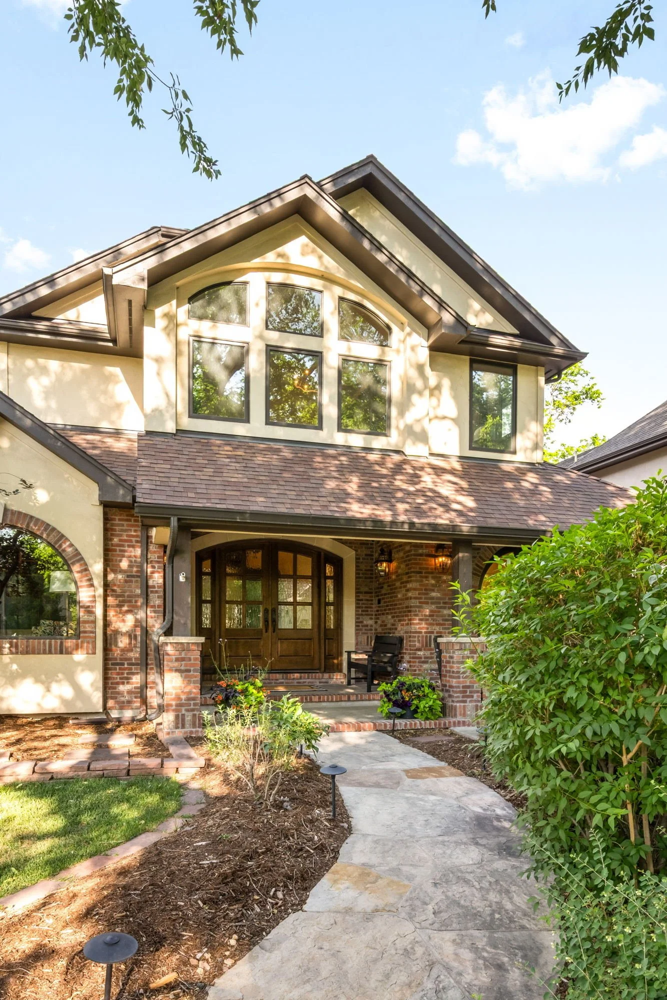 Front view of a house with a brick facade, large windows, and a welcoming porch with potted plants and outdoor seating. A stone walkway leads to the entrance, surrounded by landscaped garden with greenery and trees.