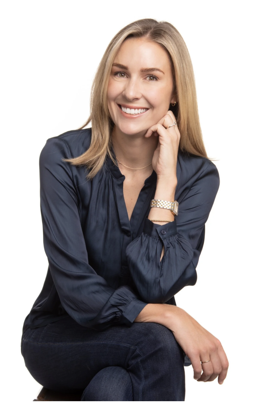 A woman with blonde hair smiling, wearing a navy blue blouse, sitting with one arm resting on her knee, against a white background.