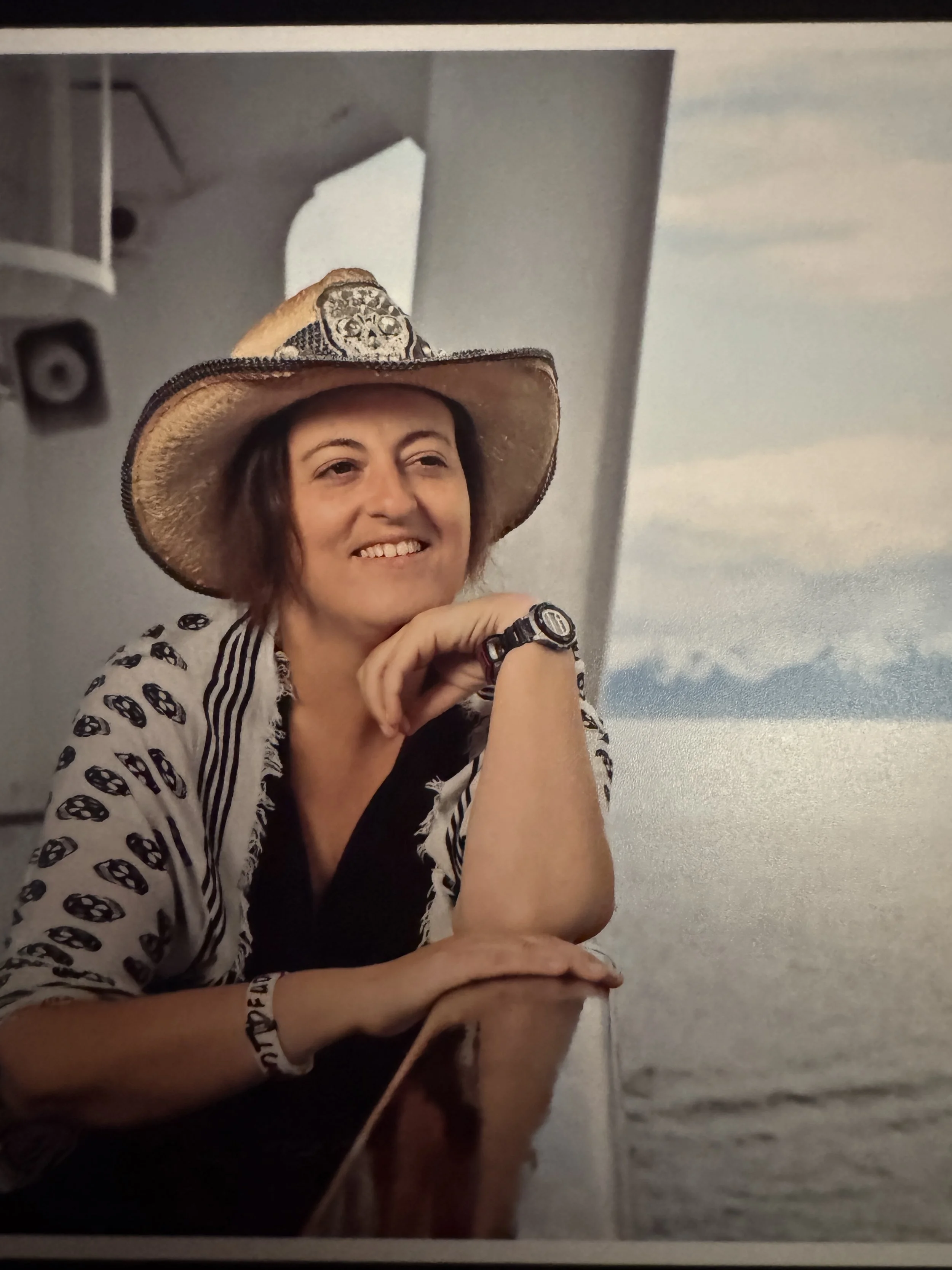 A woman smiling while wearing a cowboy hat with decorative skull design, sitting at a table with a mountain lake in the background.