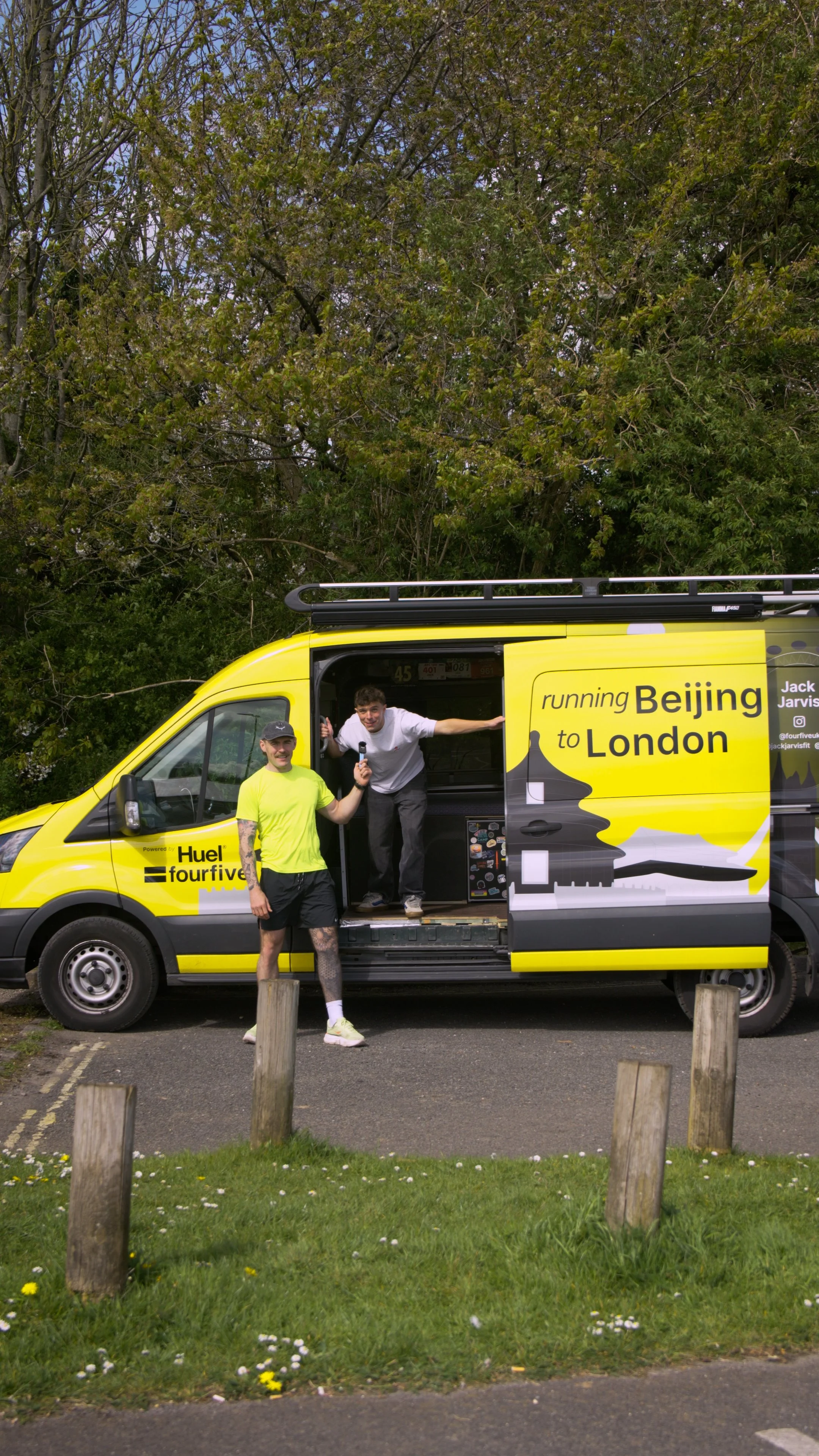 Two men stand in front of a bright yellow van with a large graphic that reads 'running Beijing to London' and 'powered by Huelfourfive.' One man, in a neon green t-shirt and black shorts with tattoos, stands outside the van, smiling. The other man, in a white shirt and black pants, leans out of the van doorway, holding a microphone and giving a thumbs-up. The van is parked outdoors with trees and a wooden fence in the background, and a grassy area with small white flowers in the foreground.