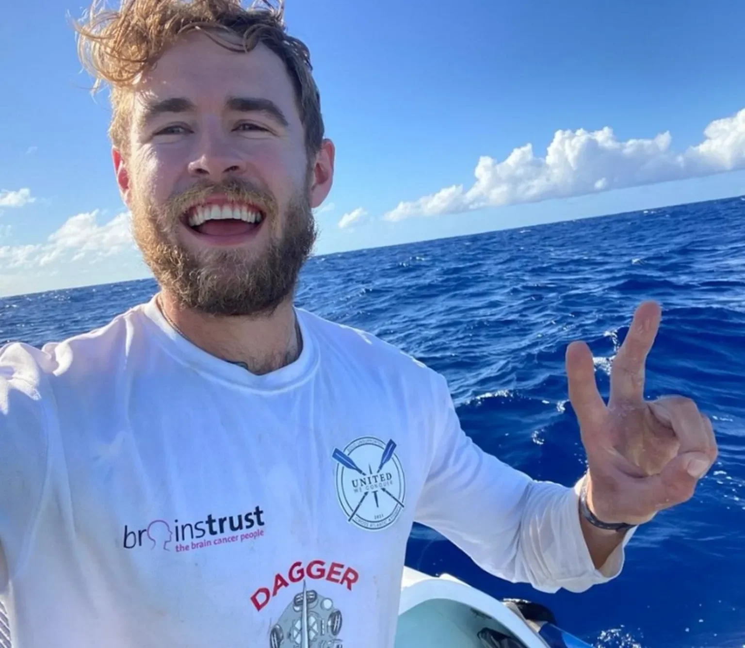 A smiling man with a beard and curly hair taking a selfie on a boat in the ocean, making a peace sign with his hand, wearing a white long sleeve shirt with logos, under a blue sky with clouds.