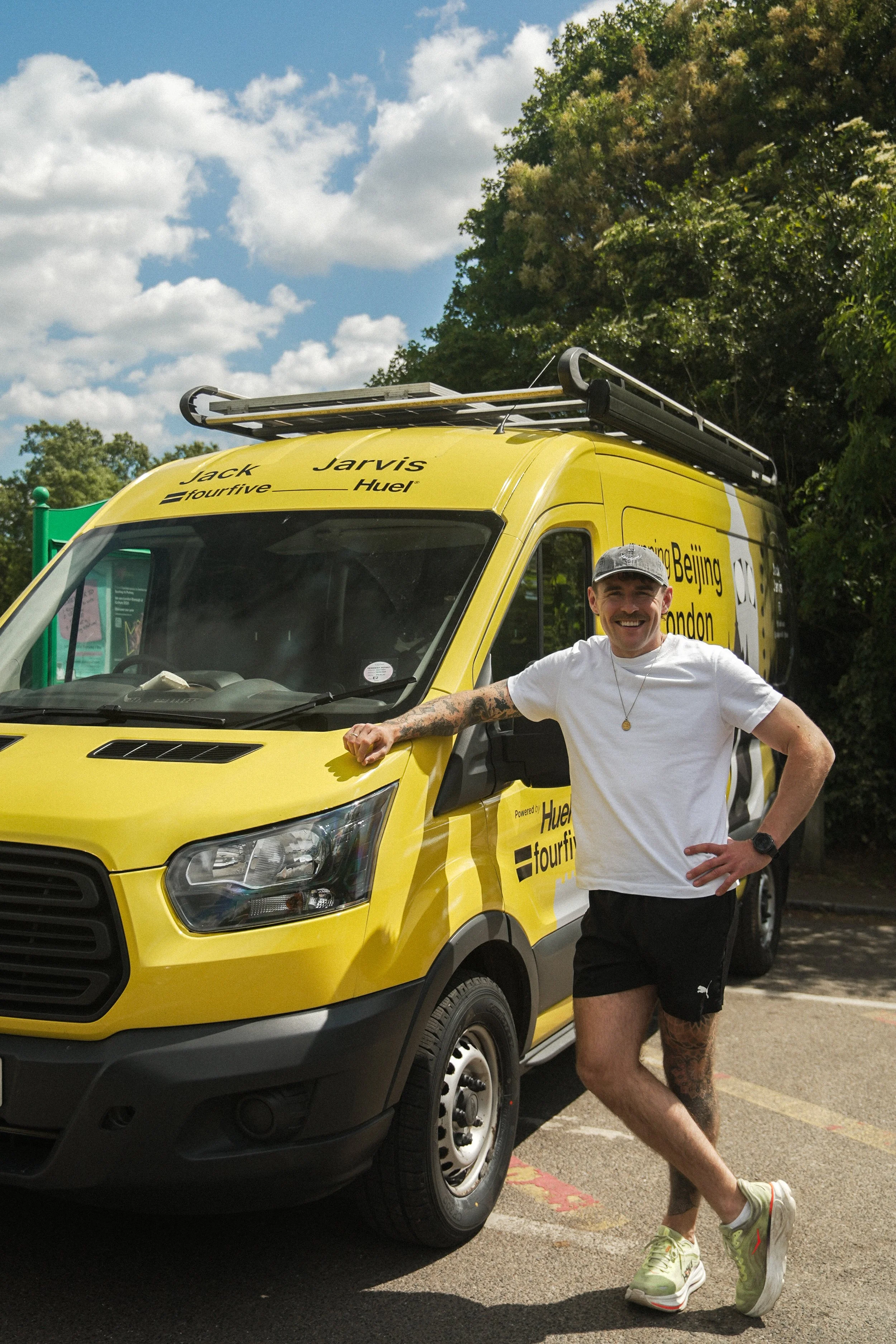 A man with tattoos on his right arm and left leg, wearing a gray baseball cap, white t-shirt, black shorts, and green Nike sneakers, standing with one hand on his hip and the other resting on a yellow van. The van has advertising for 'Fourfive' and 'Huel', with a roof rack on top. The background includes green trees and a partly cloudy sky.