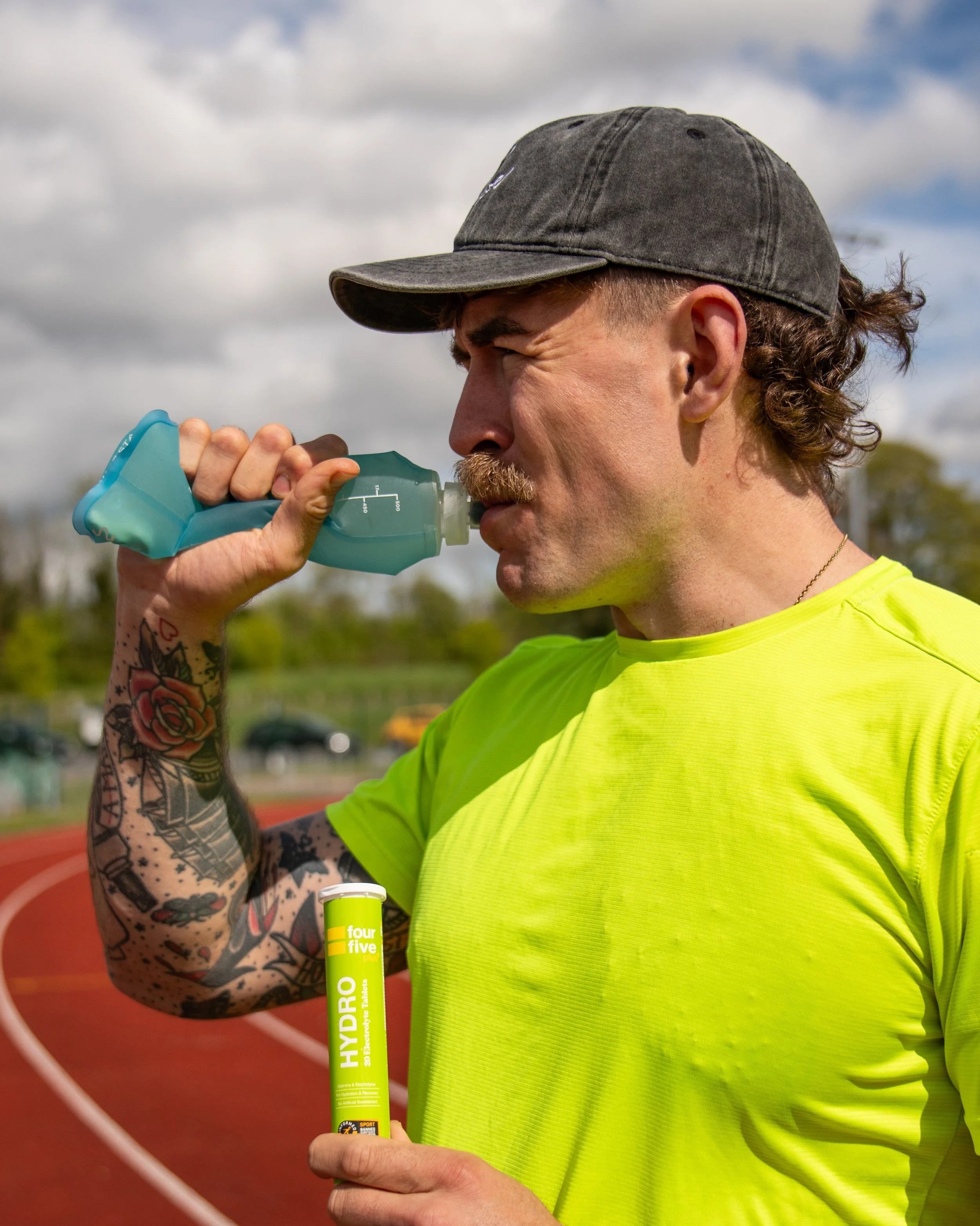 A man wearing a gray cap and a bright yellow shirt drinking from a green hydration tube at an outdoor sports track with a blue sky and clouds overhead.
