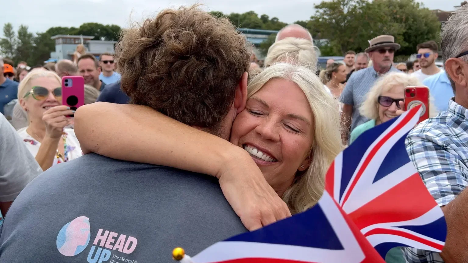 A woman with blonde hair is hugging a man with brown hair during a crowd gathering. She is smiling with her eyes closed. People around are taking photos and smiling, with some holding British flags.