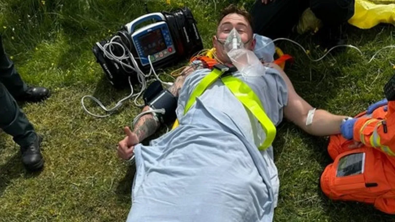 Man lying on the grass with medical equipment, giving a thumbs-up gesture while wearing a face mask and a reflective vest.