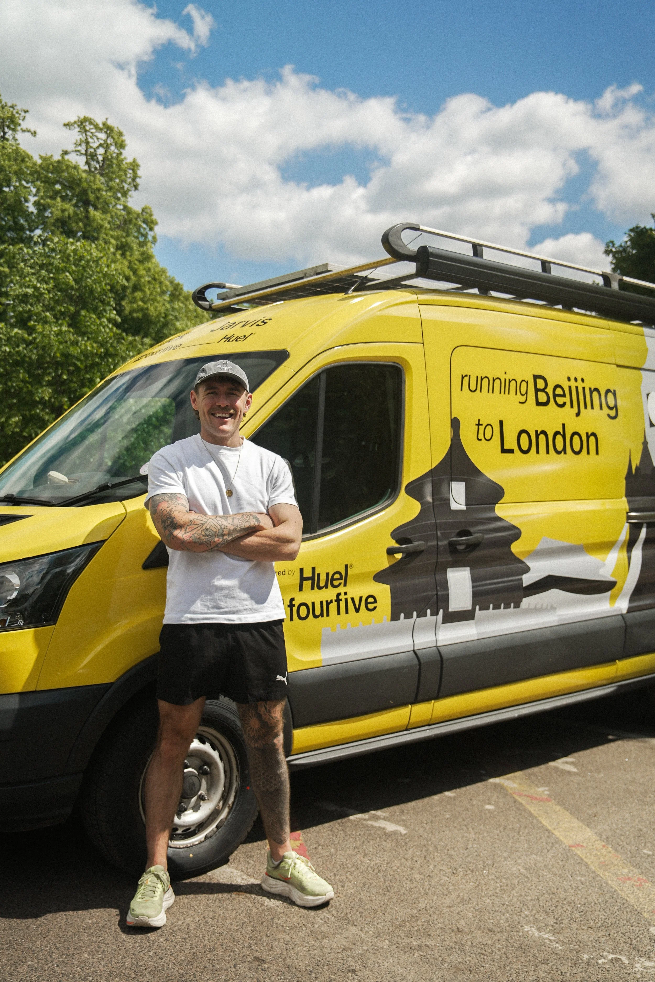 Young man with tattoos on his left arm, wearing a gray cap, white t-shirt, black shorts, and green running shoes, standing in front of a yellow van with a sign that says 'running Beijing to London.' The sky is partly cloudy, and there are trees in the background.