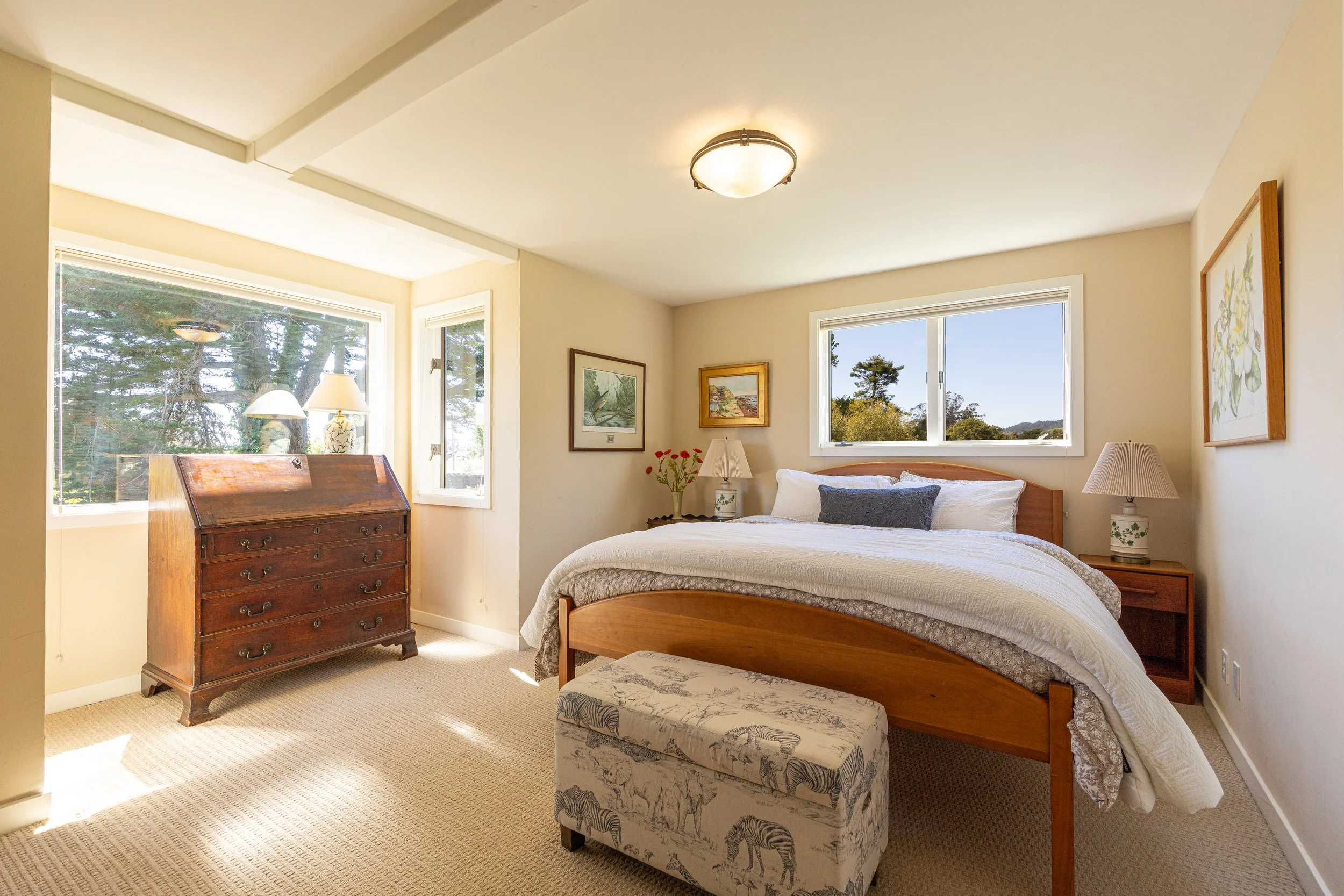 A bright bedroom with large windows showing trees outside. The room features a wooden bed with a white bedspread, matching nightstands with lamps, a cushioned ottoman with a zebra print, framed artwork on the walls, and a vintage chest of drawers. A 