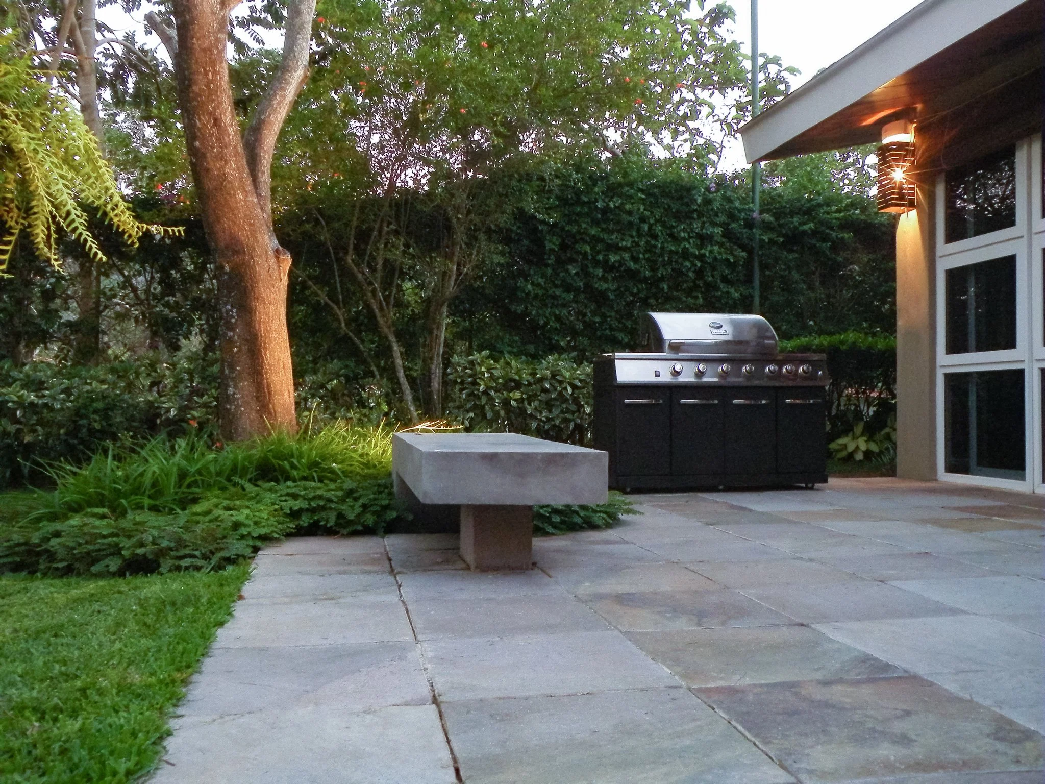 A backyard patio area with a stainless steel barbecue grill, surrounded by green plants, trees, and hedges, with a building's corner including large windows and a light fixture visible on the right side.
