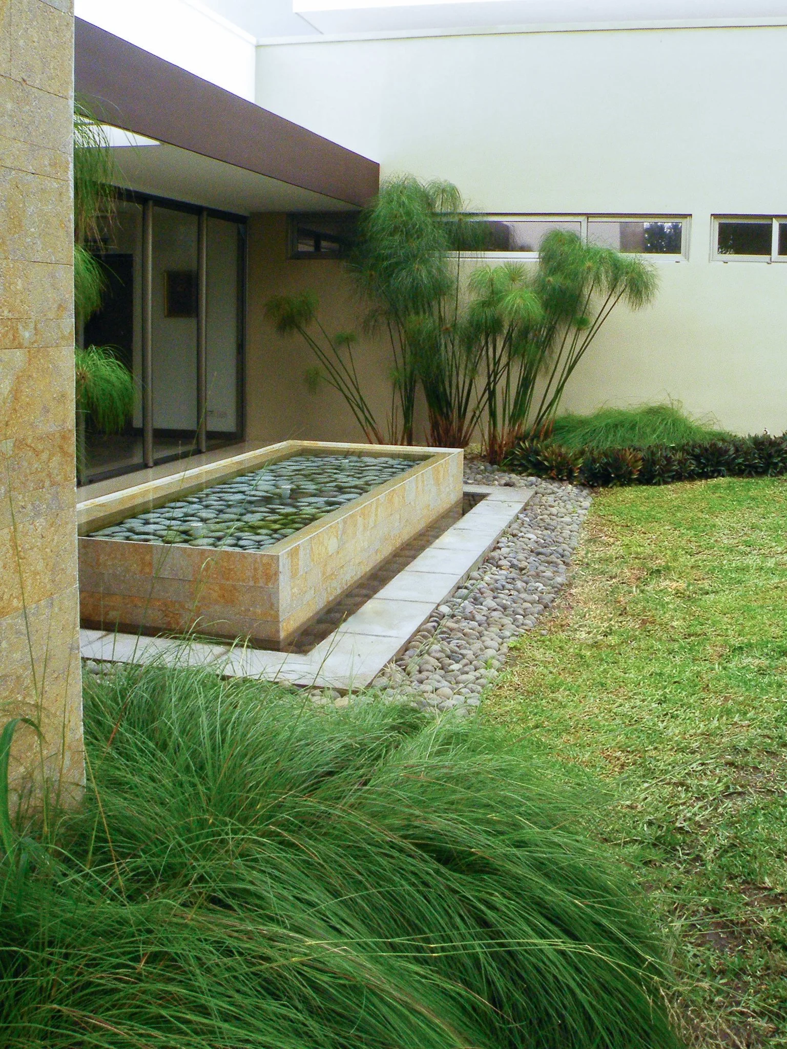 Exterior view of a modern house with a garden, featuring a water feature with rocks, planters with tall green plants, and a grassy lawn.