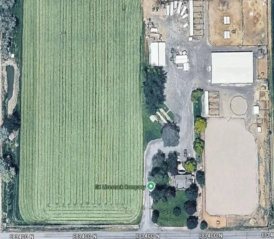 Aerial view of farmland with a large green field on the left and a facility with multiple structures, paved areas, and scattered trees on the right.