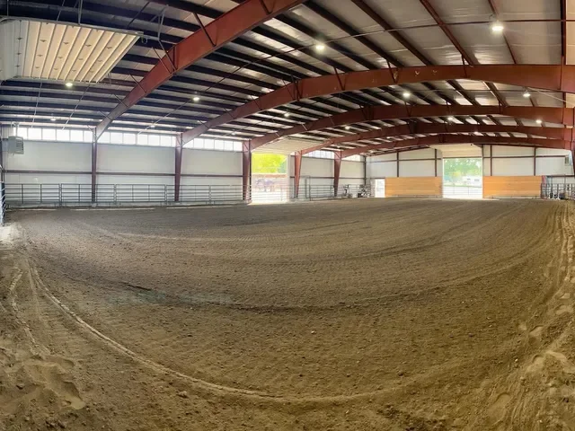 Empty indoor dirt riding arena with high ceiling and metal beams, surrounded by railings.