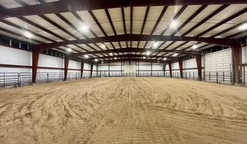 Indoor riding arena with sandy floor, metal railings, and a high ceiling with lights.