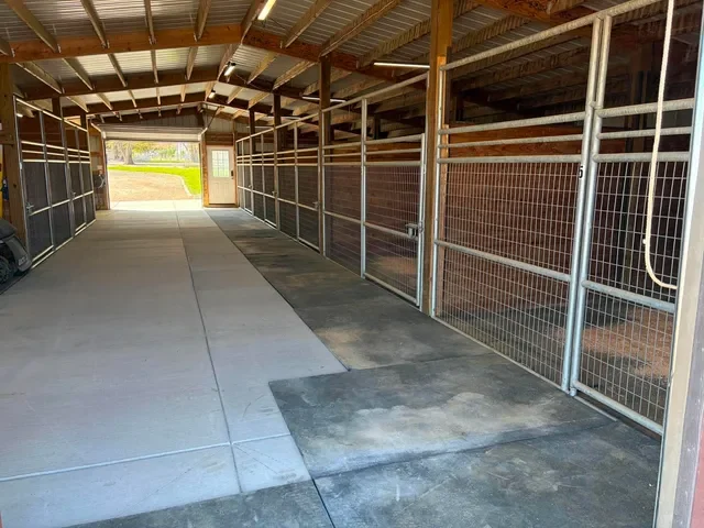 Empty horse stable aisle with metal and wood partitions on the right and a concrete and dirt floor.