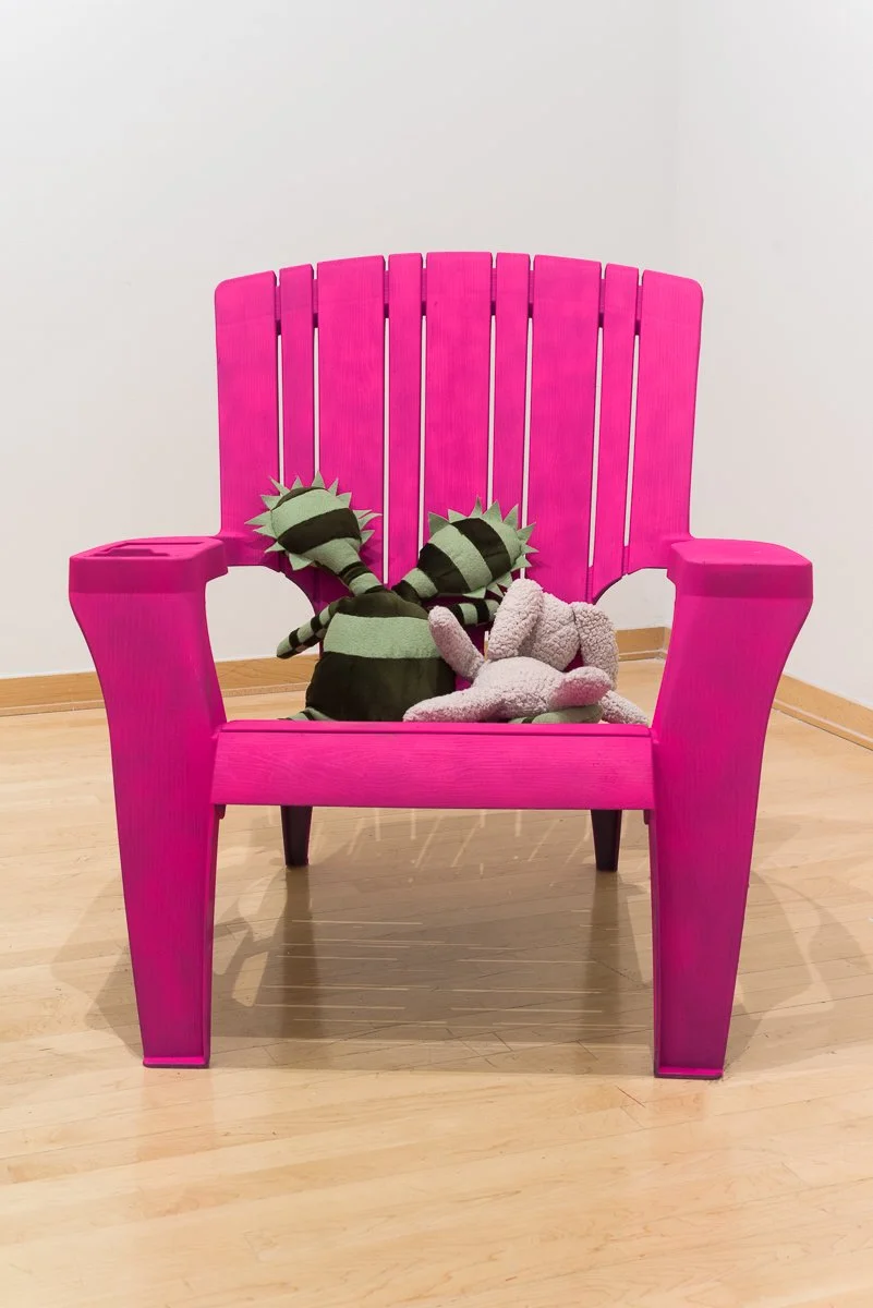 Contemporary art installation from New Mexico based artist, Sarah Heyward. Pink Adirondack chair with two plush stuffed animals sitting on it, two striped feline plush toys and a pink teddy bear, against a plain white wall and wooden floor.