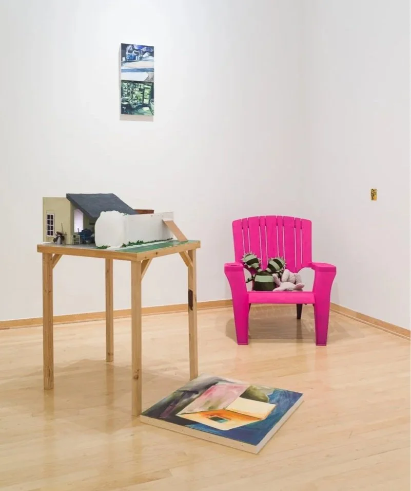 A small exhibition space with a pink Adirondack chair, a stuffed animal, a miniature house with a sloped roof on a wooden table, a colorful open book on the floor, and a painting on the wall.