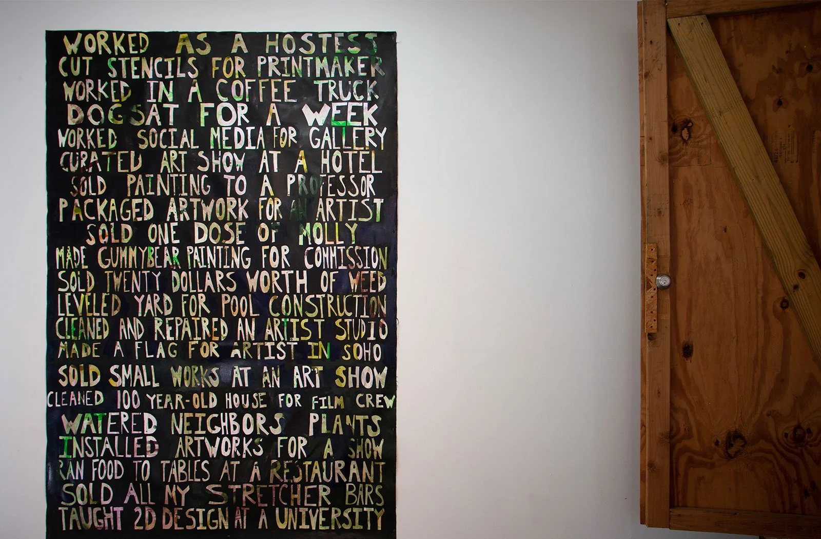 A colorful text poster on a white wall describing various artistic and creative work experiences by New Mexico-based artist and educator Sarah Heyward.
