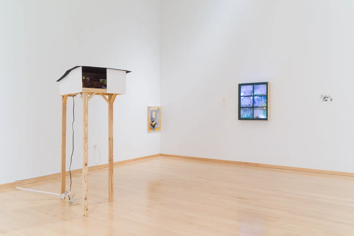 Art installation in a gallery with a house model structure on wooden legs and framed artwork on white walls.