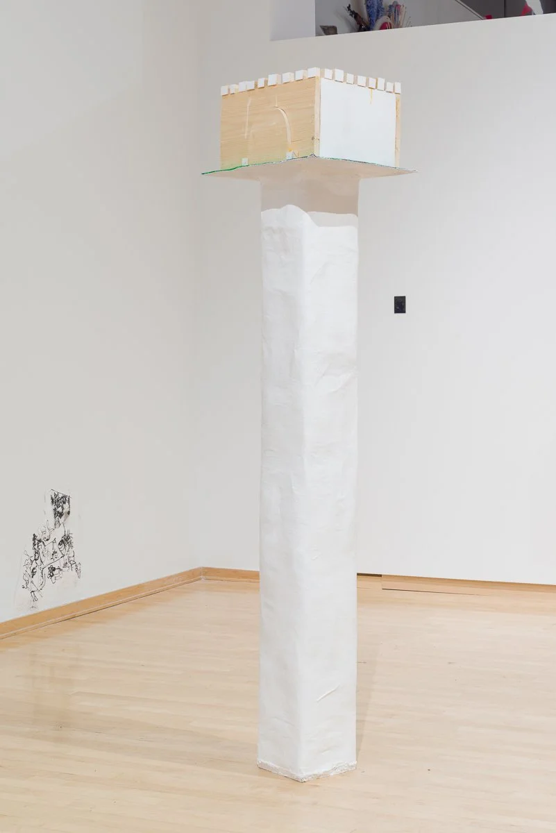 A tall art installation consisting of a white paper-covered column supporting a wooden cube with a small white block on top, set in a room with light-colored hardwood floors and plain white walls.