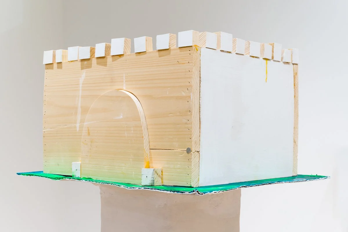 A small wooden model structure resembling a house with an arched entrance, placed on a green base.