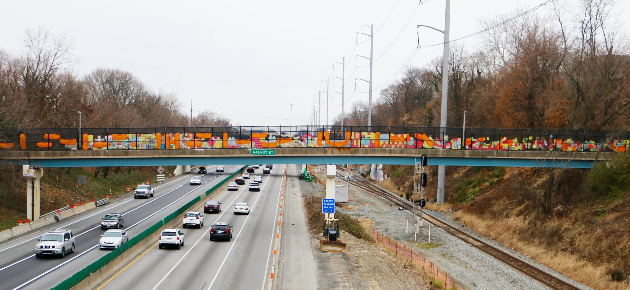 Sarah Heyward's community art project, highway with multiple cars driving underneath a bridge. The bridge has colorful decorations of recycled materials, organized by the community.