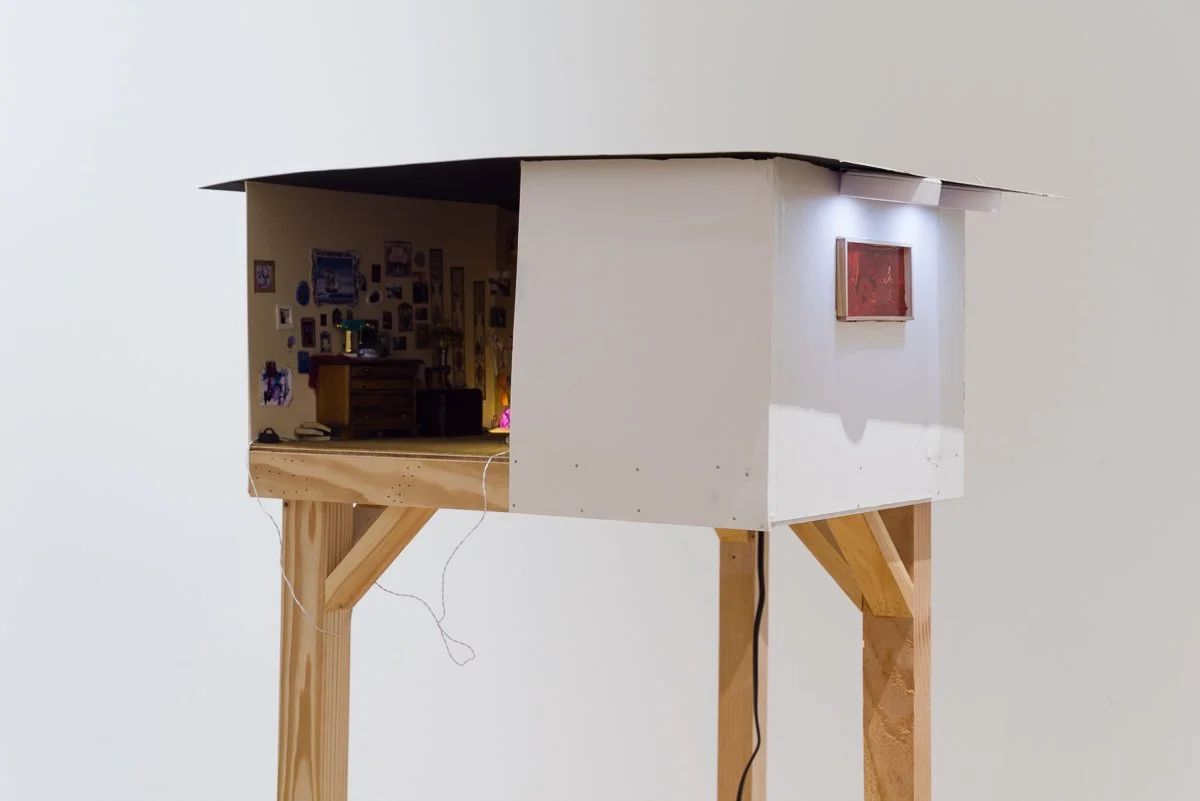 Contemporary artist and Tyler School of Art alumni Sarah Heyward's miniature house with a wooden stand, partial interior visible, and a small framed photo or painting on the exterior wall, illuminated by two small lights above it.
