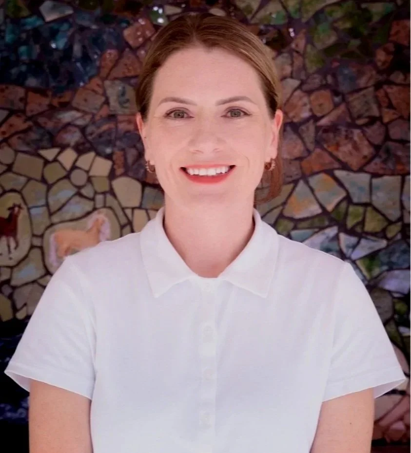A woman with short brown hair, wearing a white polo shirt, smiling in front of a colorful mural with animals and rocks.