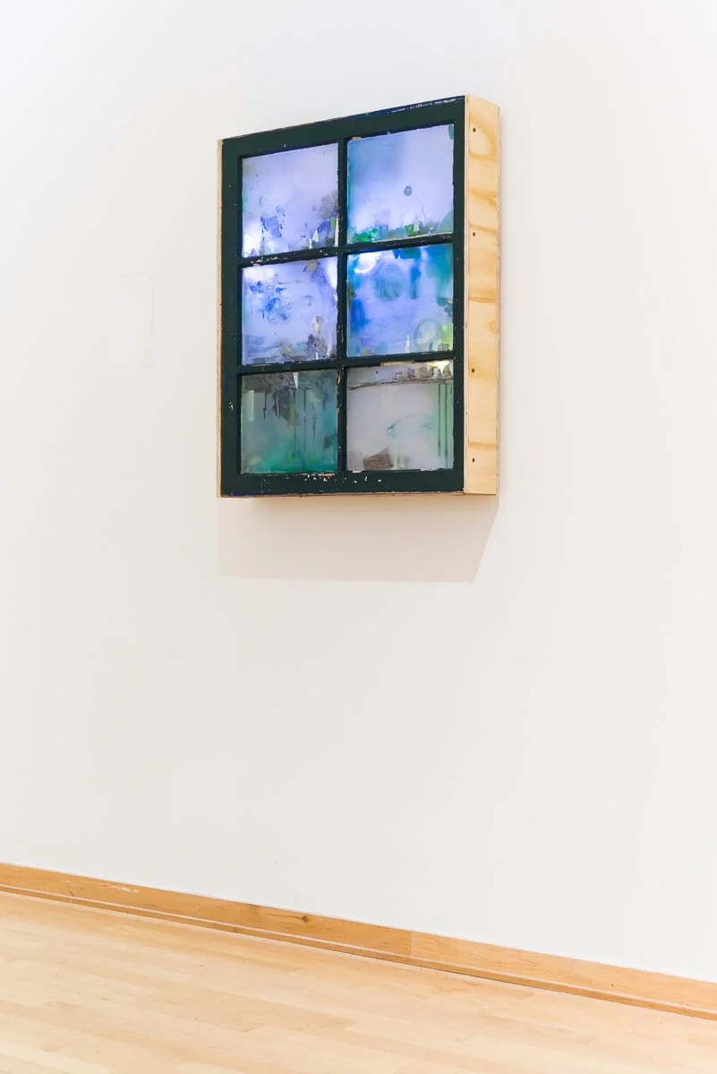 A contemporary sculptural wall piece, framed piece of art resembling a window with six divided glass panes, painted with abstract blue, green, and white colors, mounted on a white wall with a light wooden floor below.